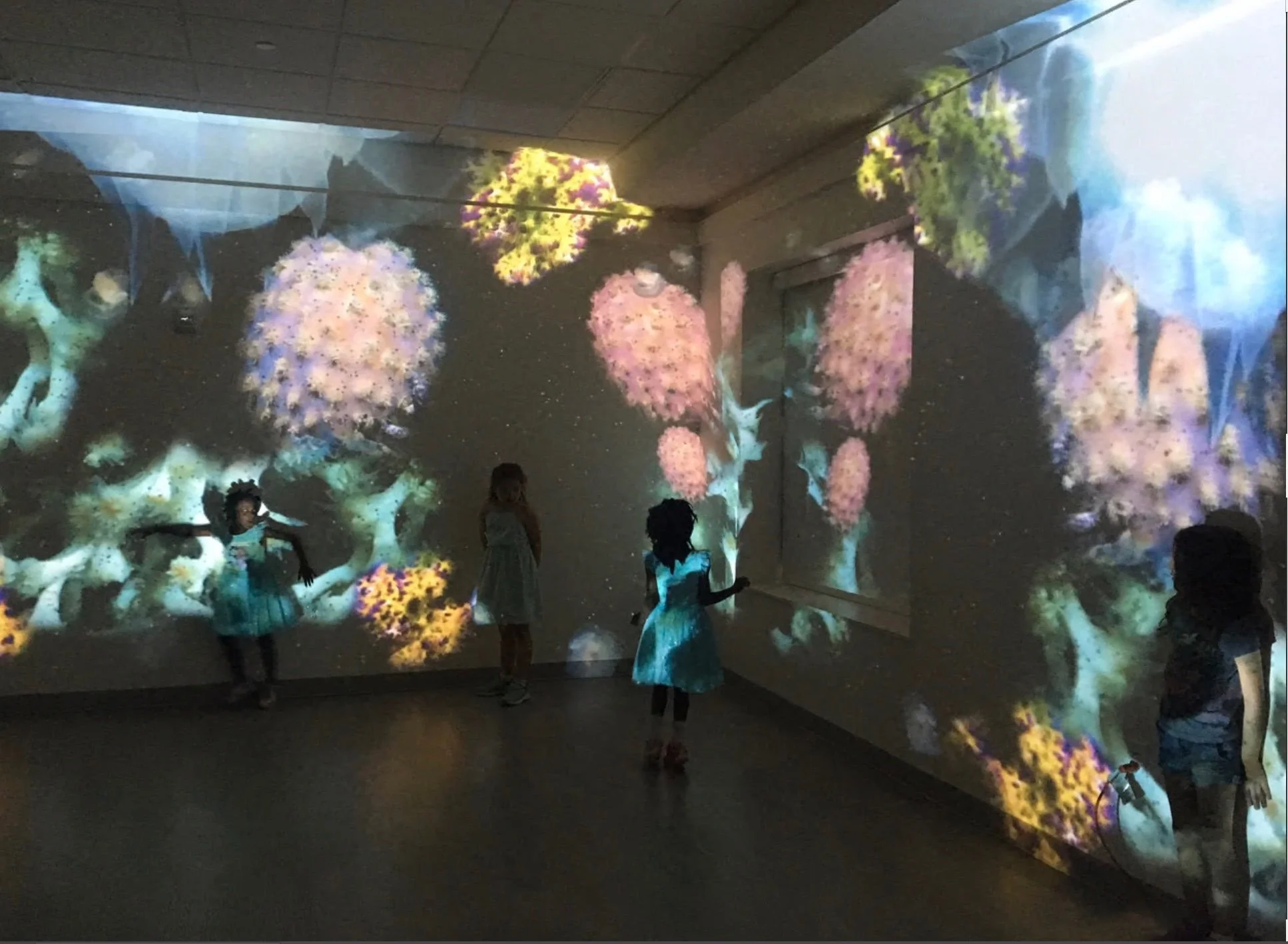 Children standing inside an immersive projection space, where abstract, organic light forms cover the walls and floor. The scene illustrates perception-led engagement, where sensory experience shapes awareness, intention,.