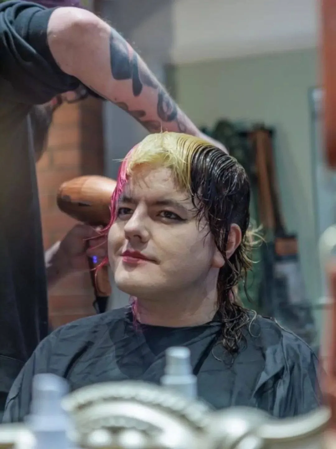 Person sitting in a salon chair getting their hair dyed with streaks of blonde and pink, while their hairdresser applies dye on top of their black hair.