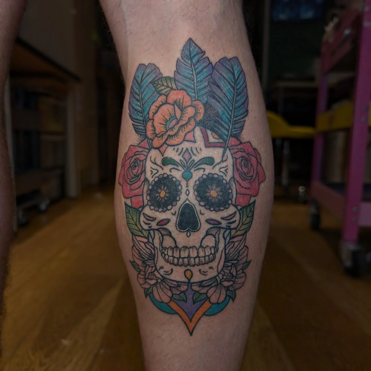 Tattoo of a decorated sugar skull surrounded by flowers and leaves on a person's leg.