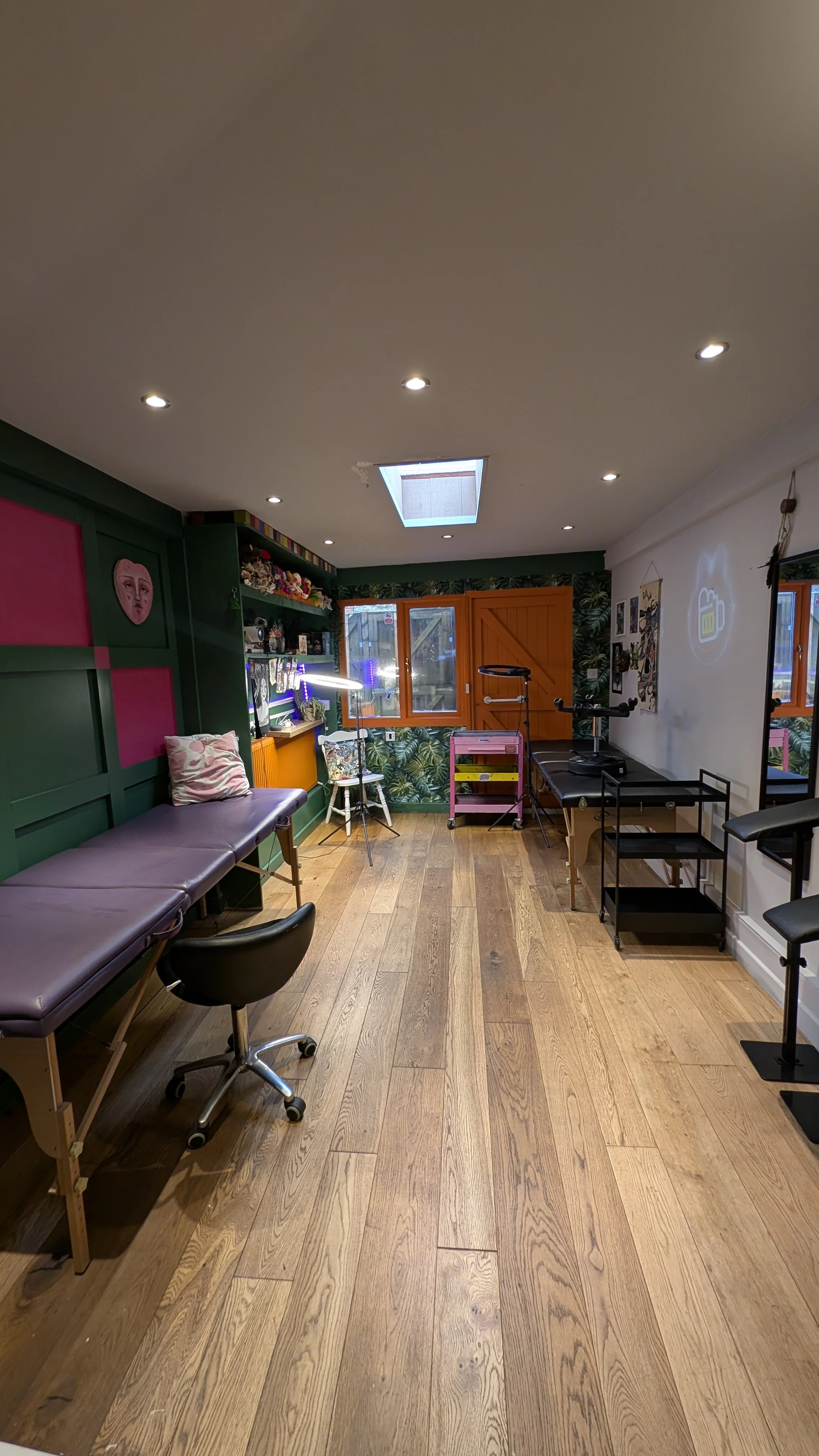 A bright room with wooden floors, a purple examination table, and a black office chair. The room has green walls with pink and face art, shelves with decorations, a window, and a ceiling skylight.