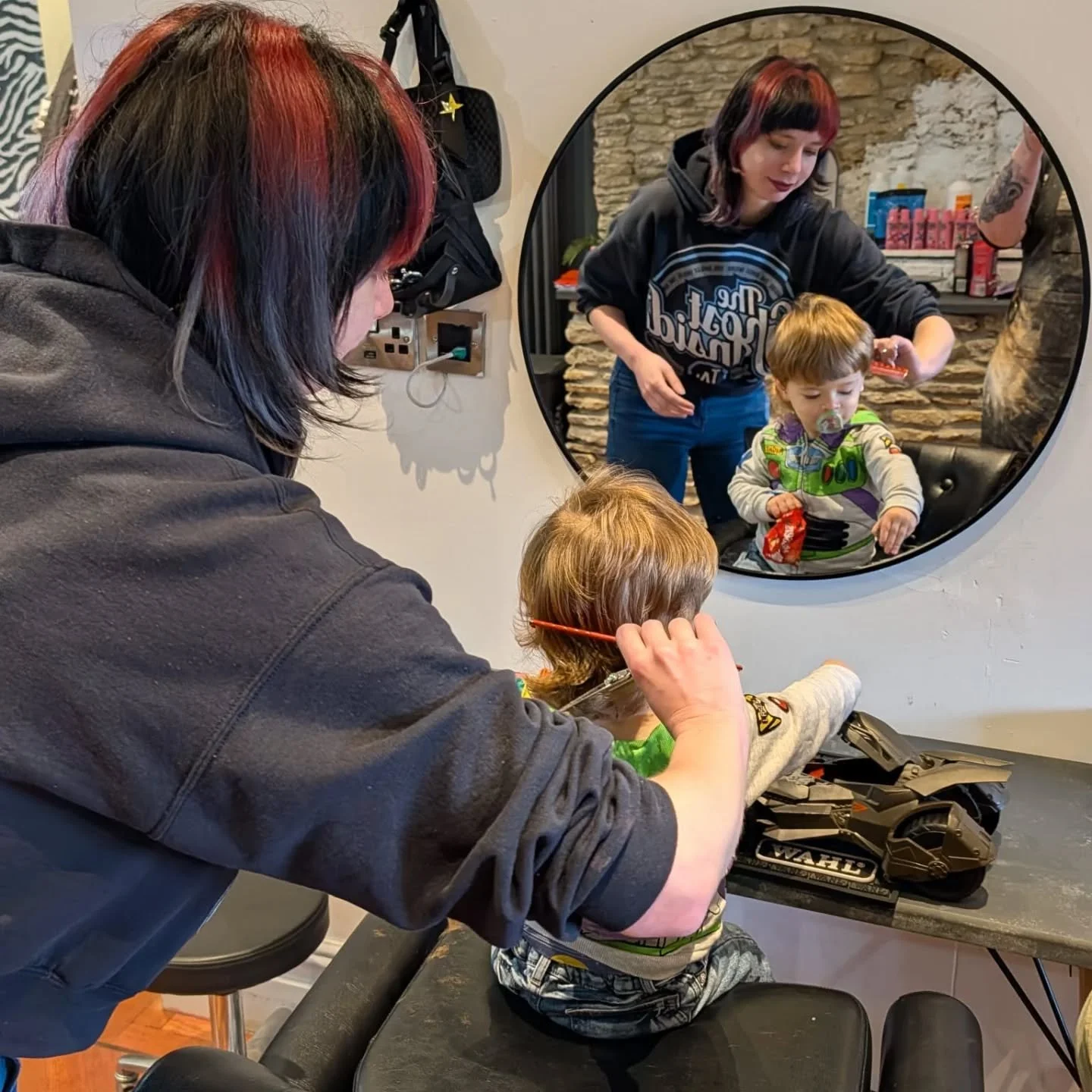 A woman with red and black hair is giving a haircut to a young boy with light brown hair who is sitting in a chair, while a woman with dark hair and a black hoodie watches through a circular mirror. The boy has a pacifier and a snack in his hand. The room has a stone wall in the background.
