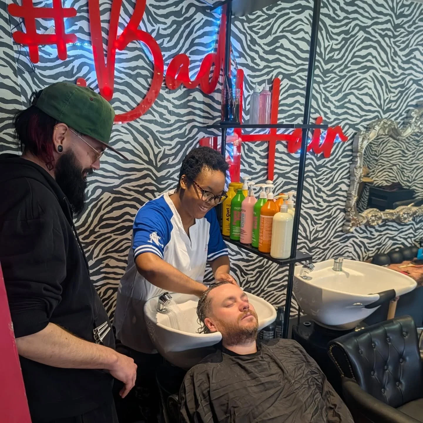 A person receiving a haircut while their hair is washed at a salon with zebra print walls, a mirror, shelves with shampoo bottles, and two hair washing sinks. There are two staff members, one washing the client's hair and the other observing.