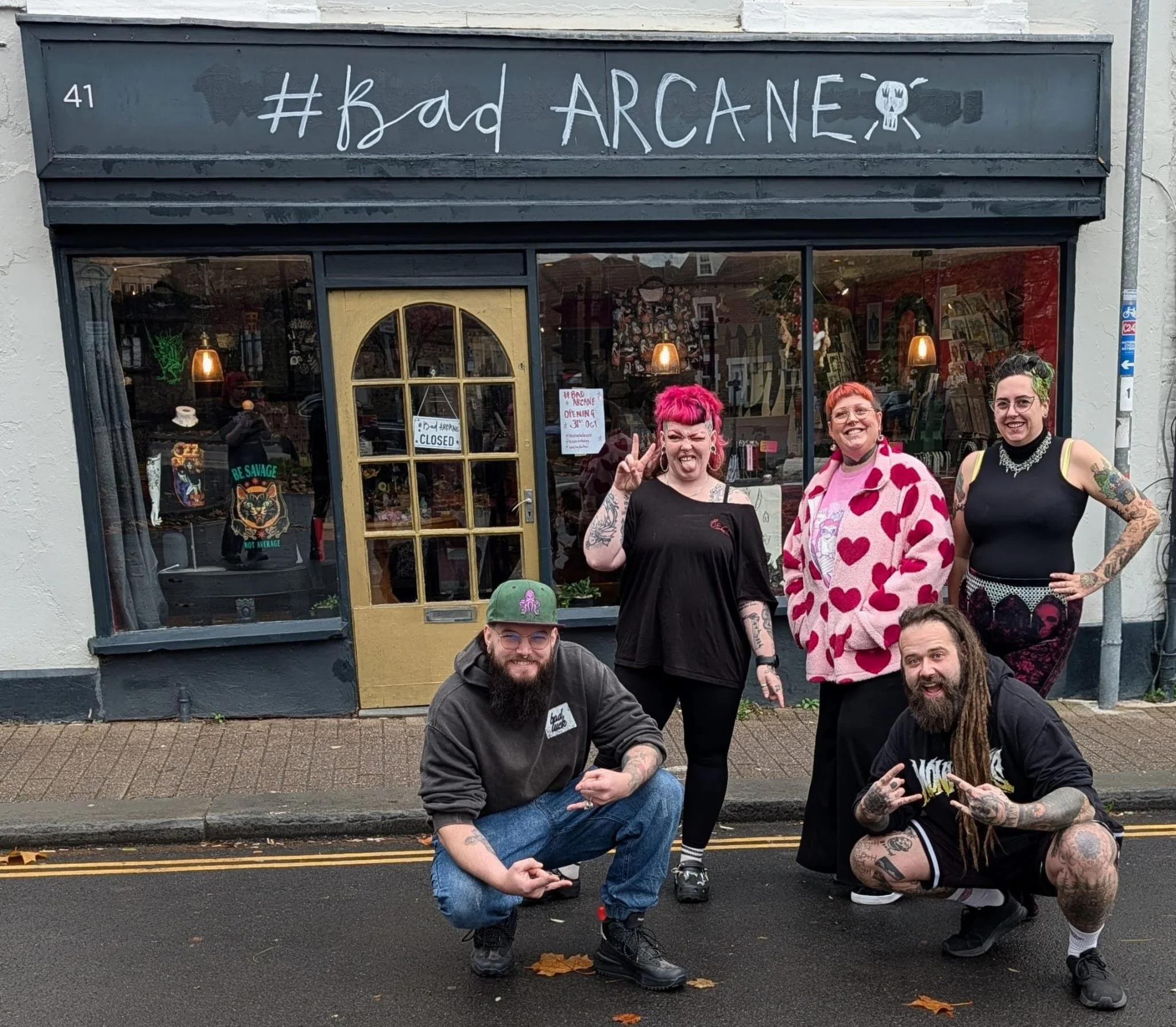Five people posing in front of a storefront named '#Bad ARCANE'. The group includes two men squatting, one with a beard and glasses, and the other with dreadlocks and tattoos. The three women are standing, one with short pink hair and making a peace sign, another with short red hair and a heart-patterned jacket, and the third with green hair and tattoos, wearing a black top with lace. The street is wet with fallen leaves.