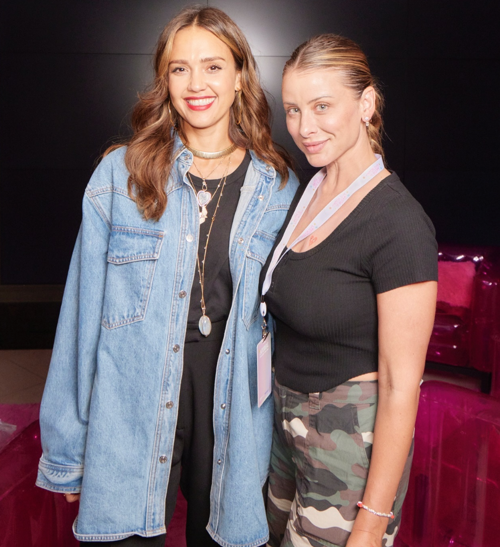 Two women standing close together, smiling at the camera. One wears a denim jacket over a black outfit with layered necklaces, and the other wears a black shirt with camouflage pants and a lanyard around her neck.