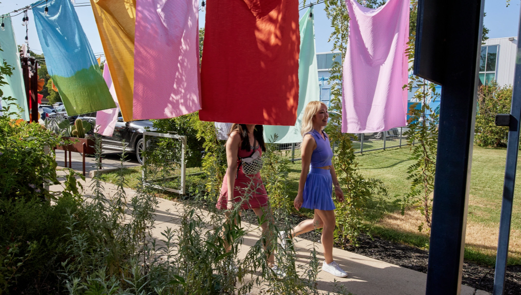 Two women walking on a sidewalk in an outdoor area with colorful laundry hanging above them on clotheslines.