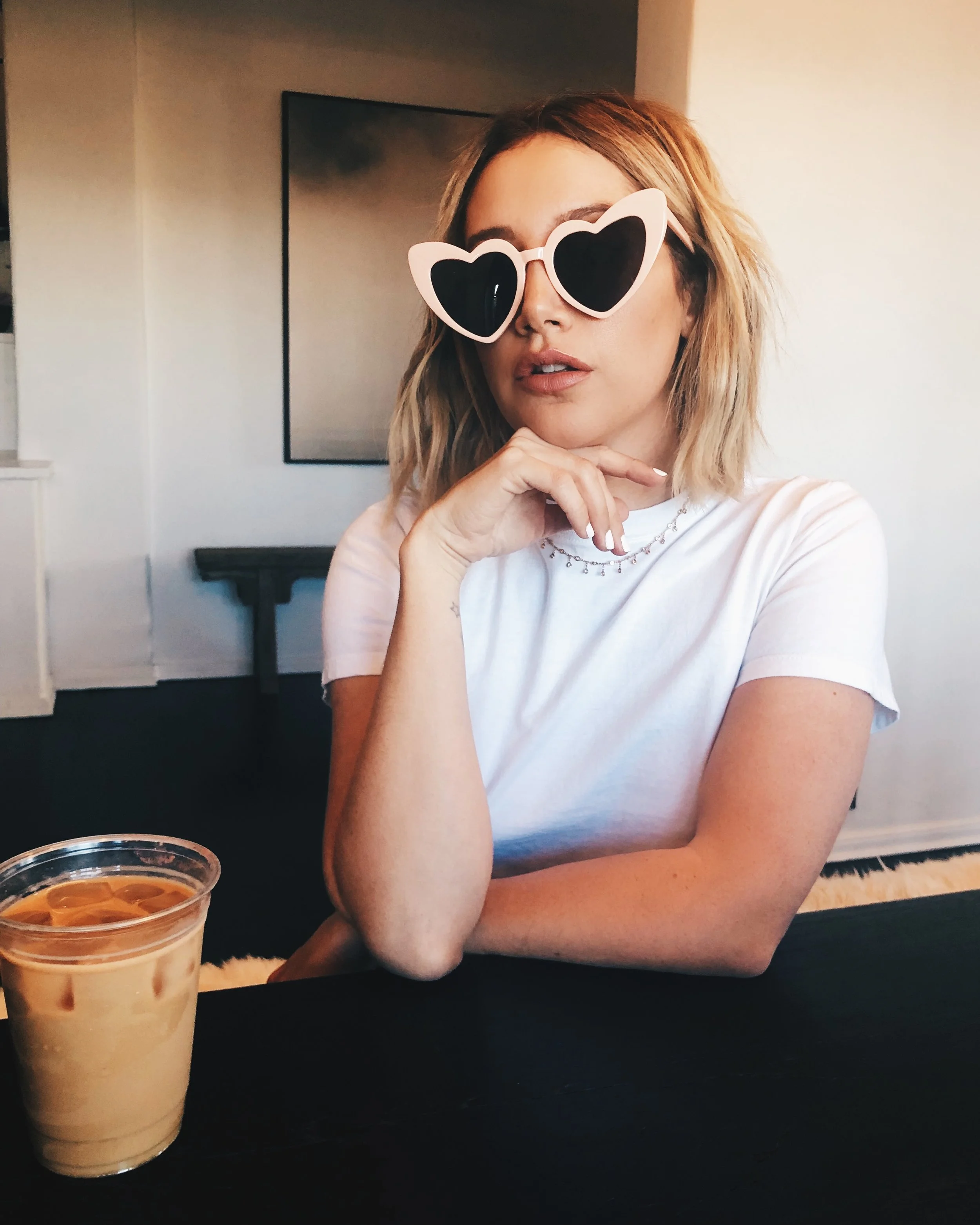 A woman with shoulder-length blonde hair wearing large, heart-shaped sunglasses and a white t-shirt sits at a table with an iced coffee in a plastic cup. She has a silver necklace and poses with her hand under her chin.