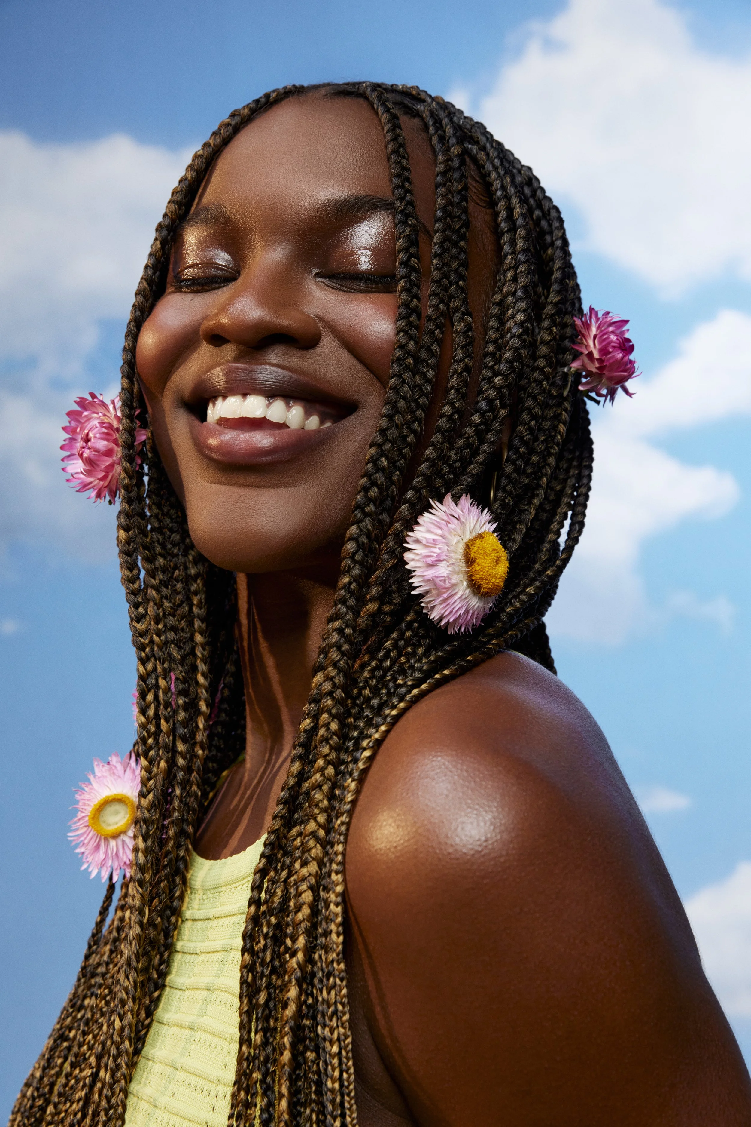 A smiling Black woman with braided hair decorated with pink and yellow flowers, standing outdoors against a blue sky with clouds.
