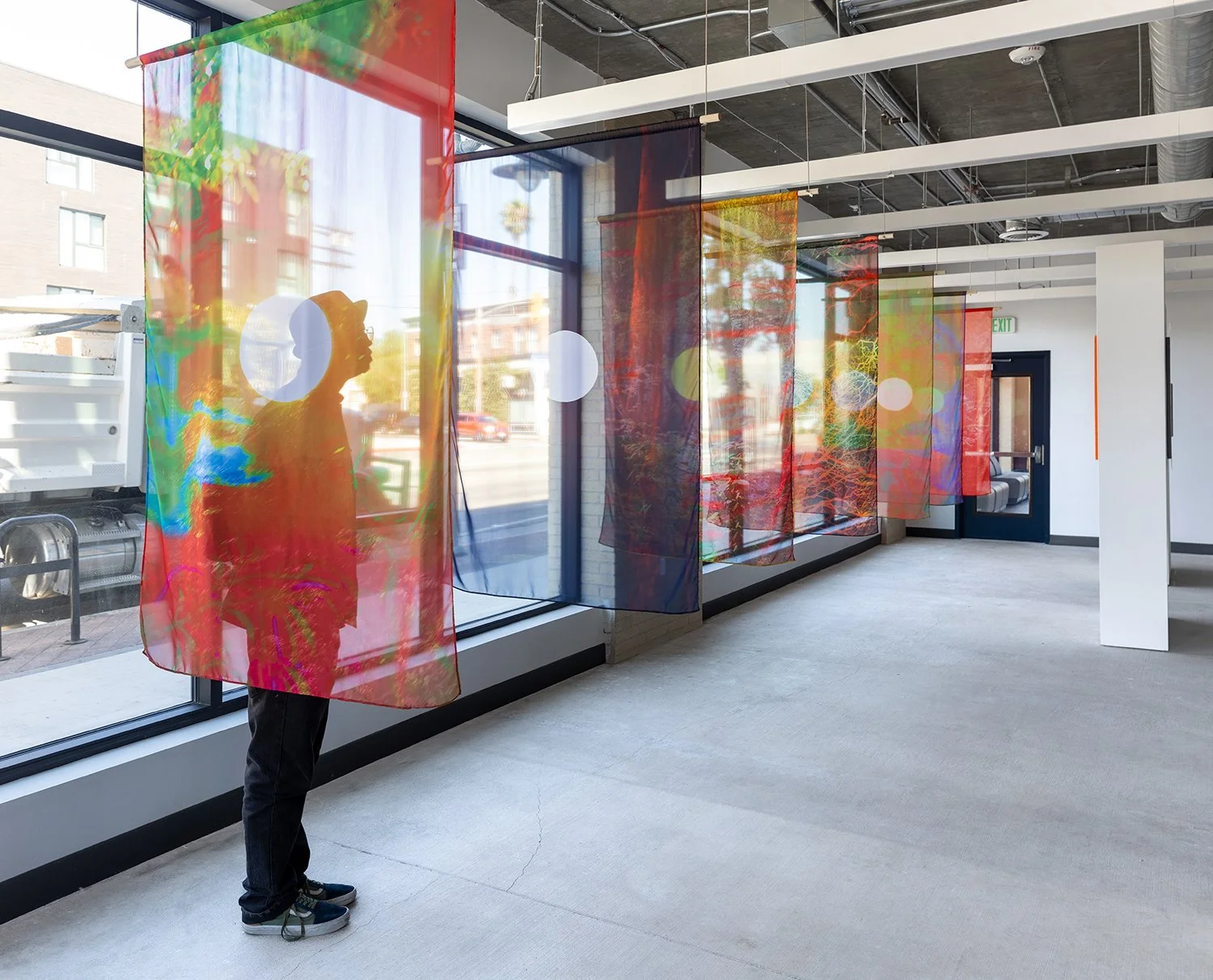 Interior of an art gallery with colorful, semi-transparent hanging fabric panels, large windows allowing natural light, and a person inspecting the artwork.