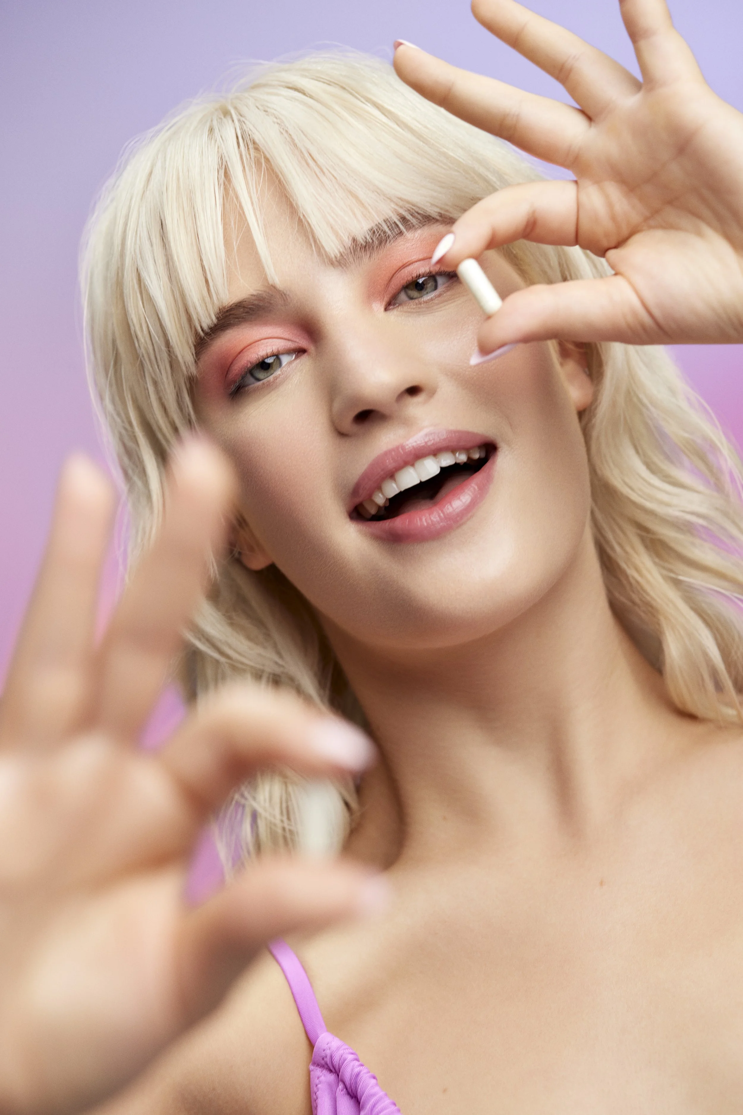 A woman with blonde hair and pink eyeshadow holding a white capsule pill close to her face, smiling, with a purple background.