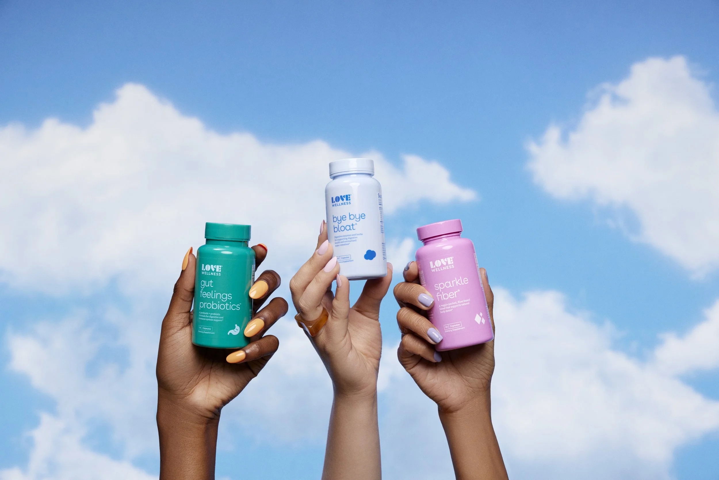 Three hands holding colorful wellness supplement bottles against a blue sky with clouds.