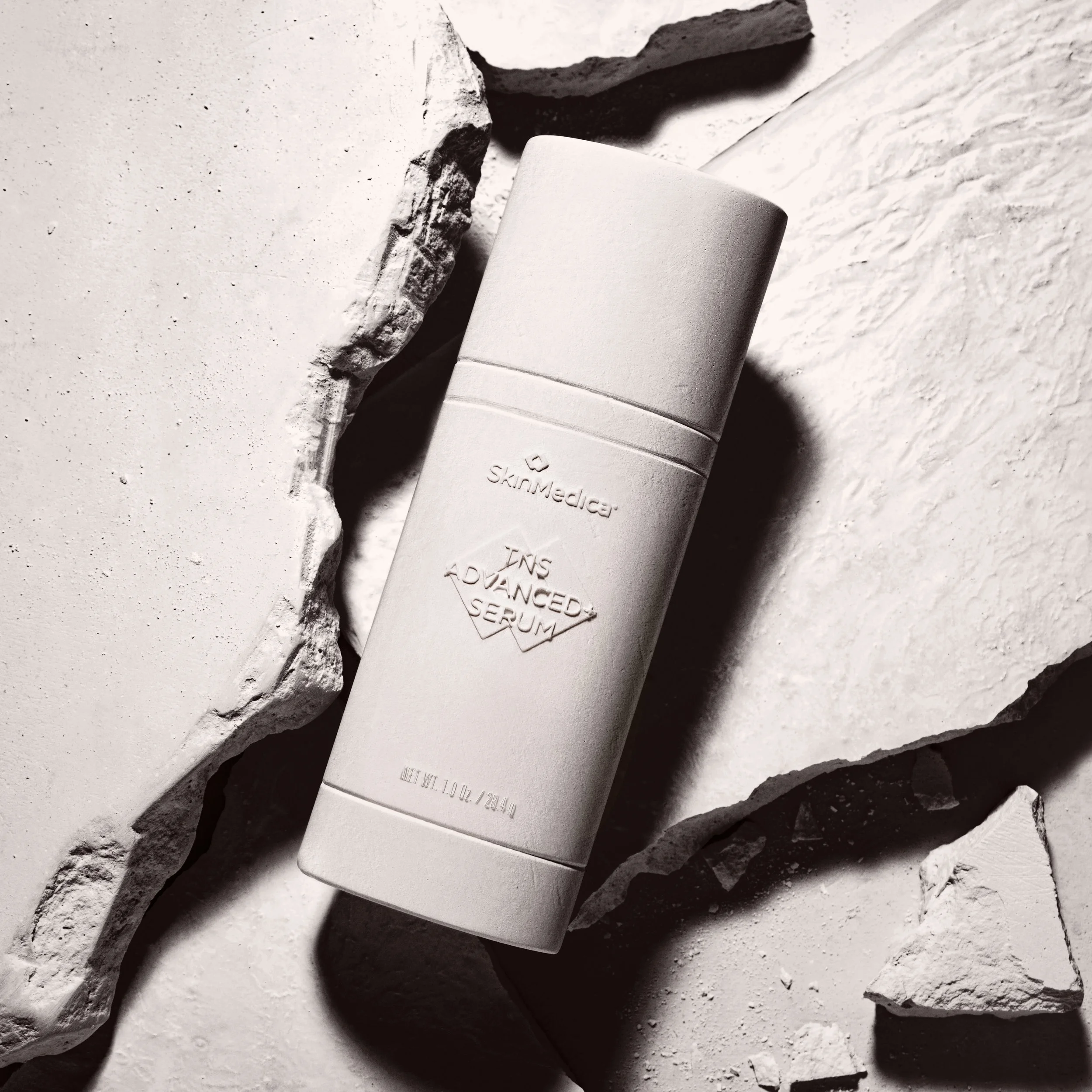 A bottle of SkinMedica TNS Advanced Serum placed on white rocks in a black and white photograph.