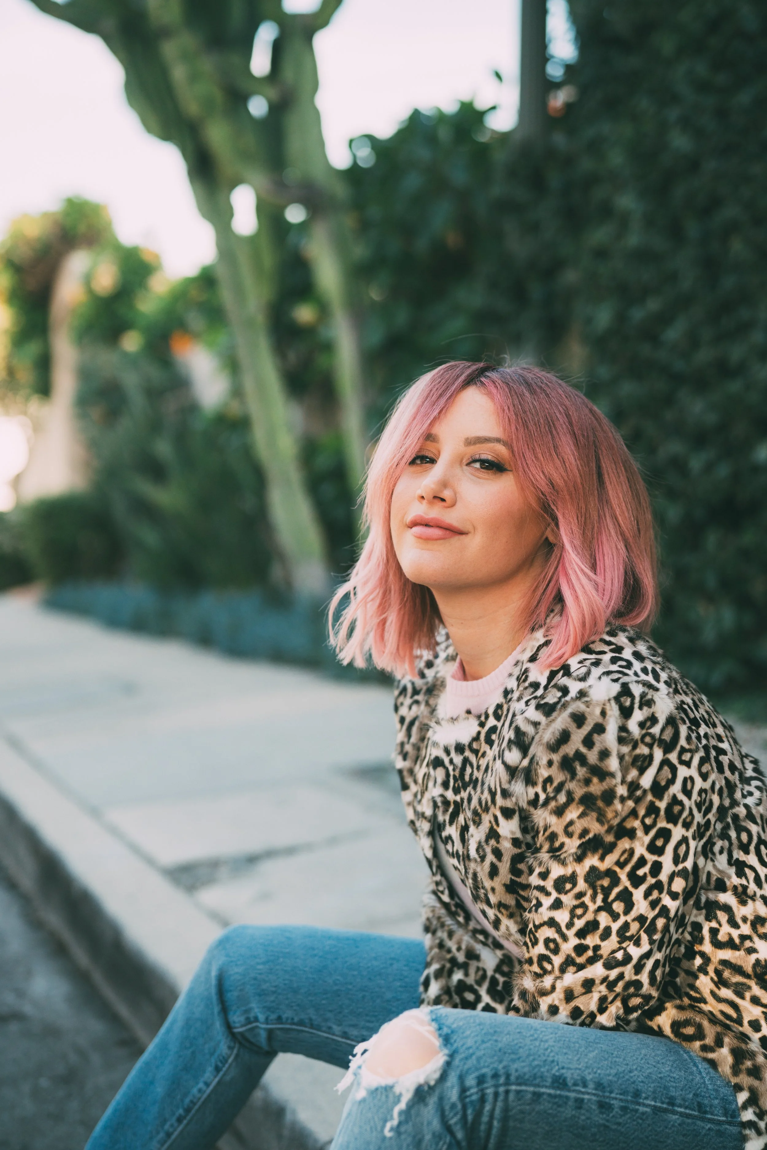 A woman with pink hair and a leopard print jacket sitting on a sidewalk with greenery in the background.