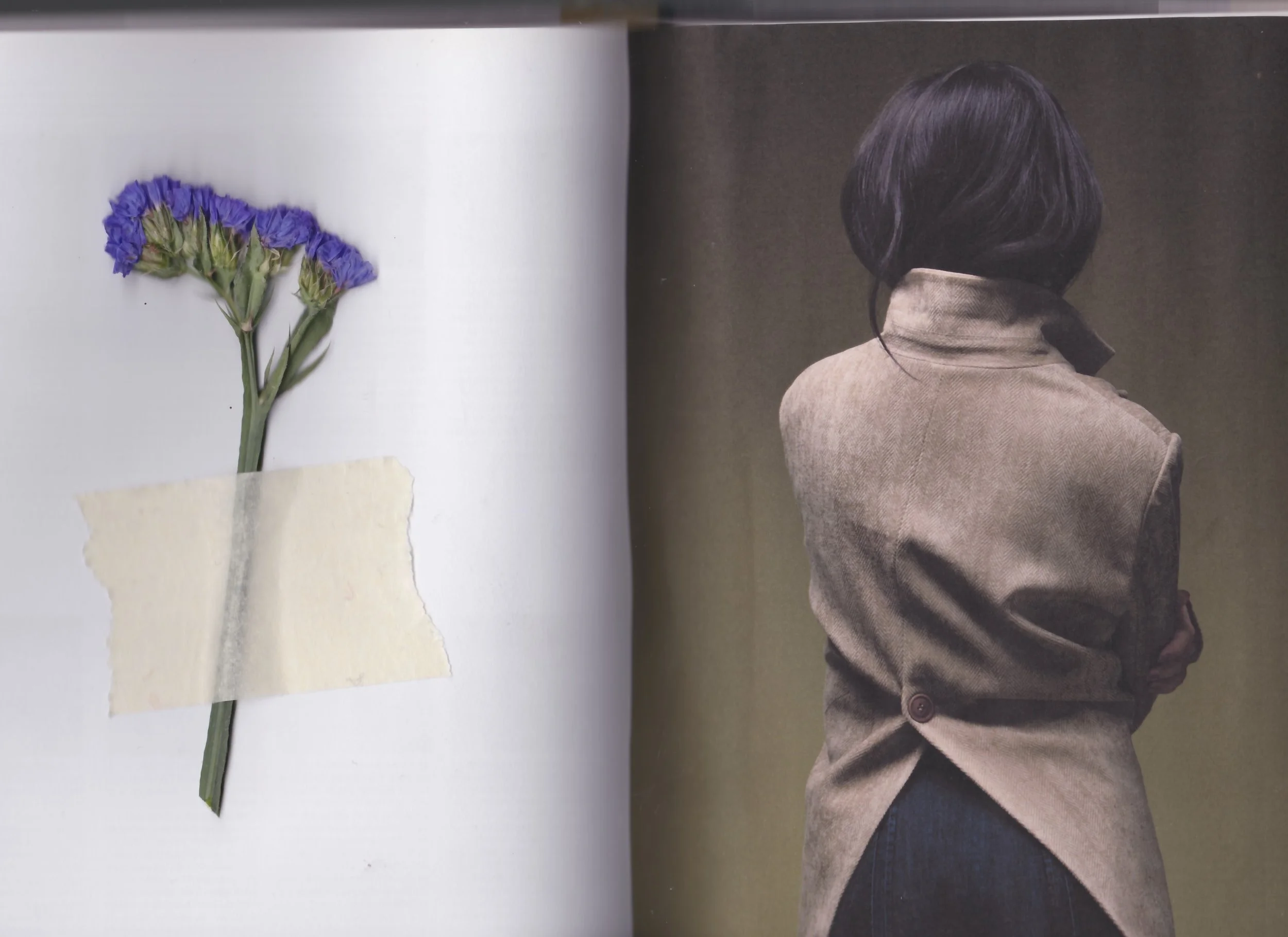 A photograph of a bunch of purple statice flowers taped to a white background and a woman with short dark hair wearing a beige coat with her back turn to the camera, standing against a plain brown background.