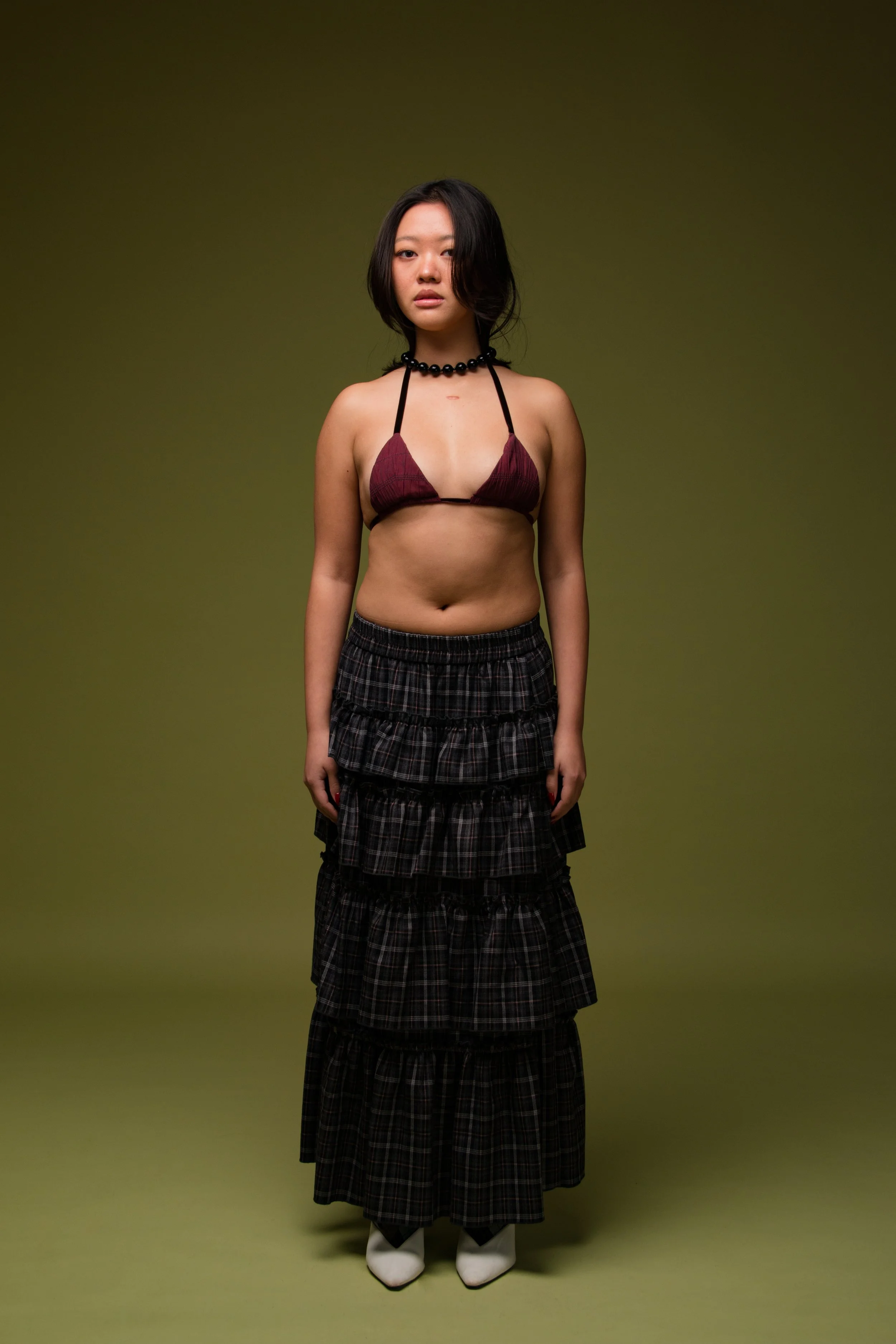 A young woman with shoulder-length dark hair stands against a plain green background, wearing a maroon bikini top, a black beaded necklace, a layered black plaid skirt, and white ankle boots.