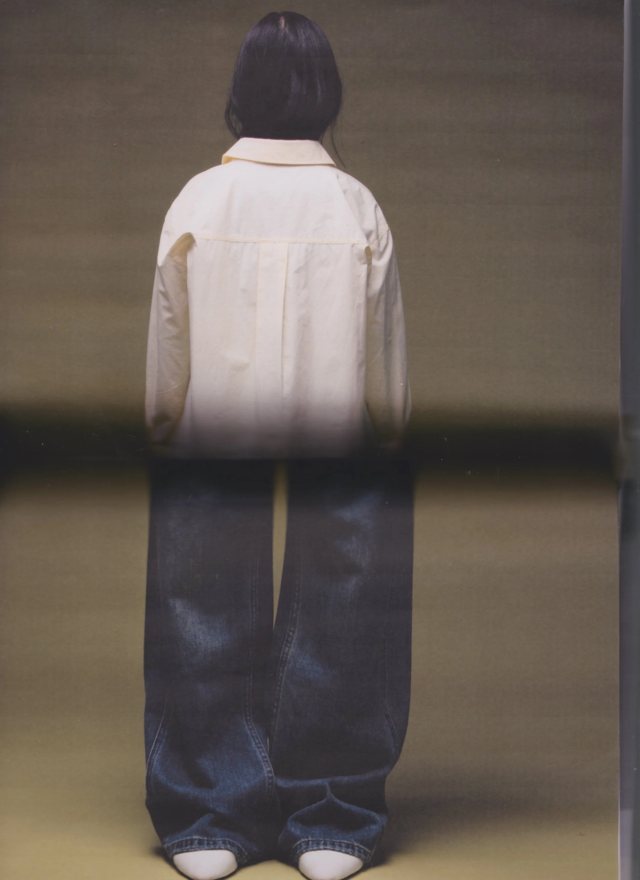 A person with dark hair wearing a cream-colored jacket and baggy dark blue jeans, facing away from the camera. The person is standing against a plain background.