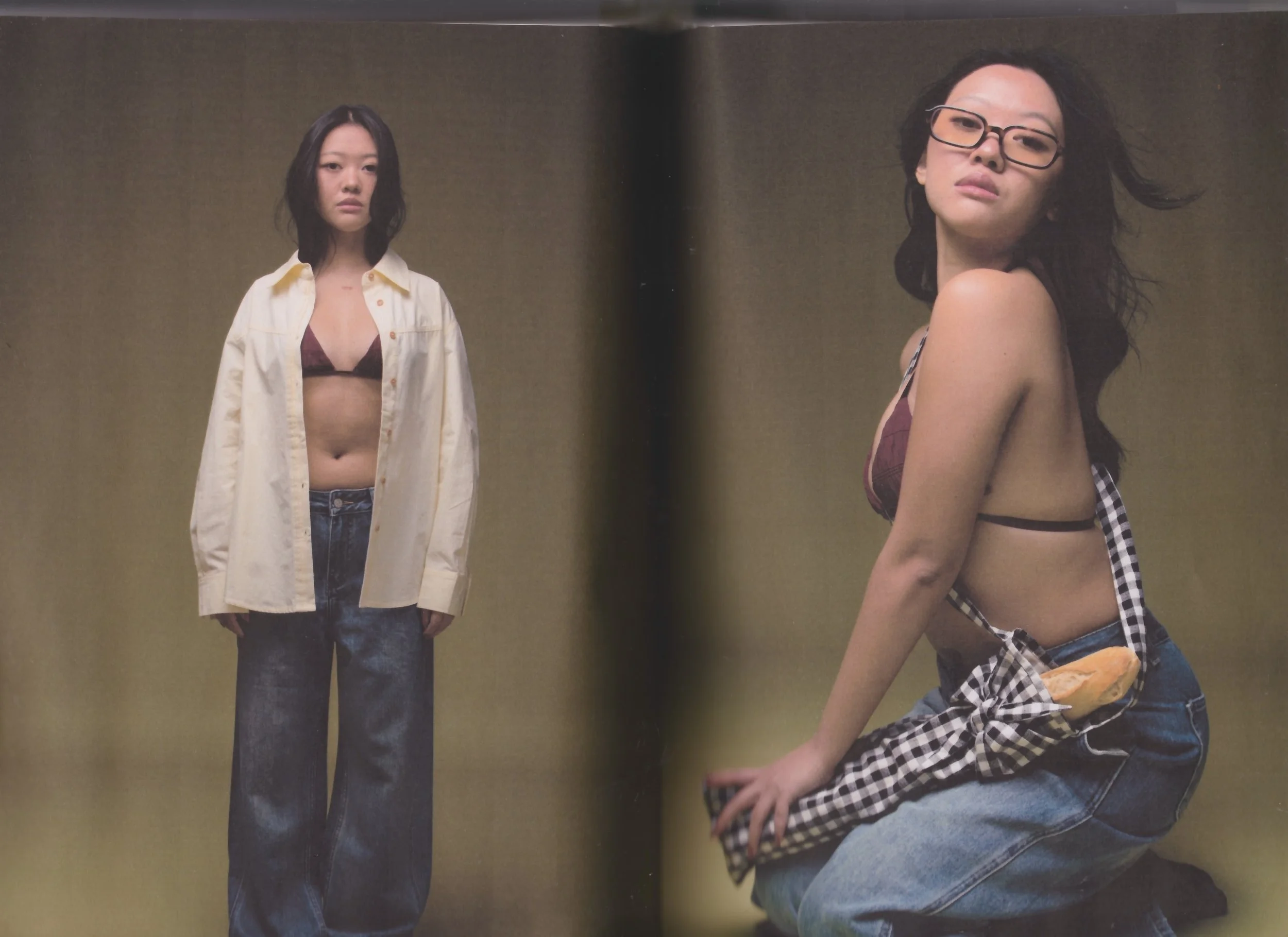 Two women in front of a plain backdrop. The woman on the left is standing, wearing a beige oversized shirt over a bikini top and jeans. The woman on the right is kneeling, looking at the camera, wearing glasses, a bikini top, and jeans, with a piece of clothing tied around her waist.