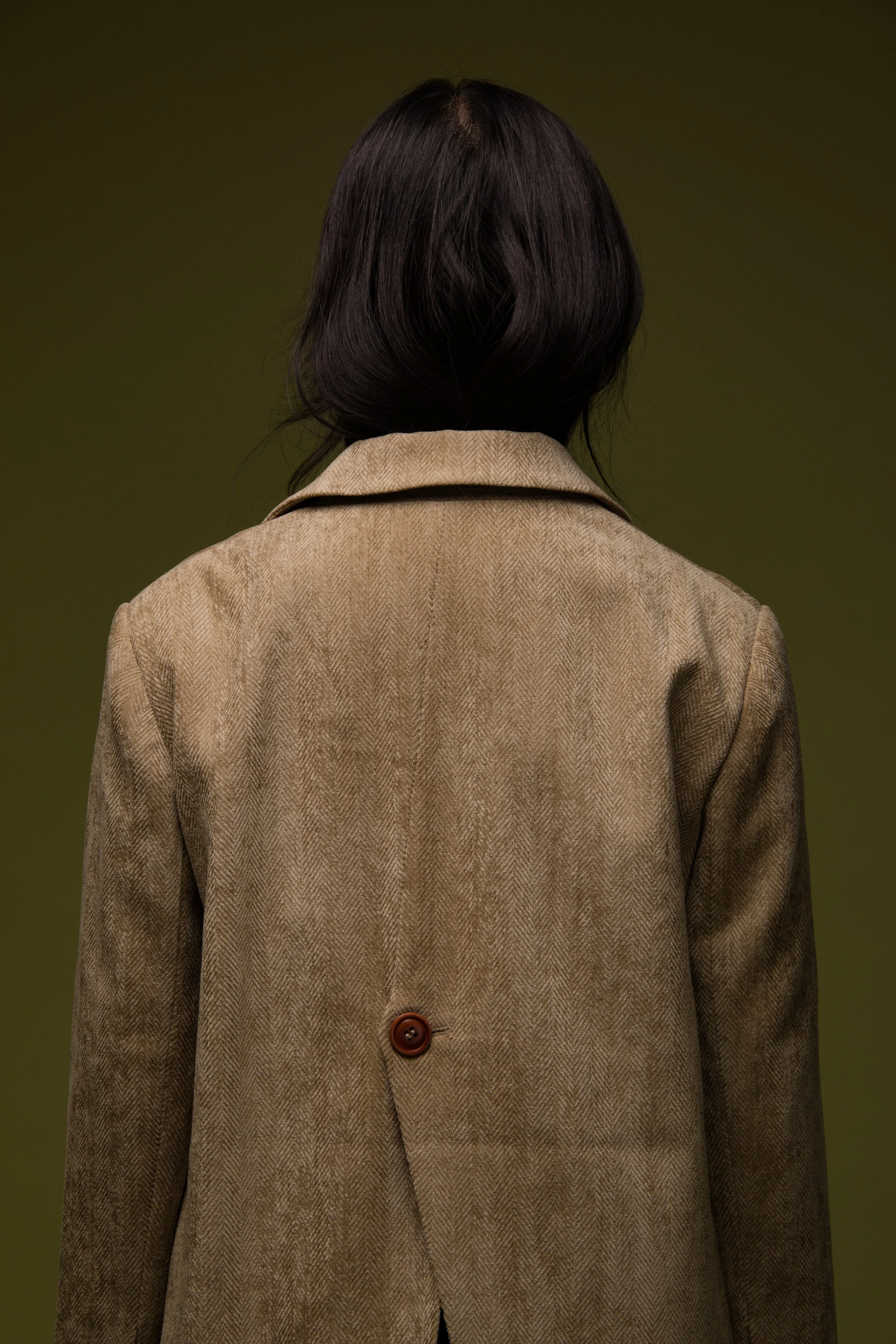 Back of a person with shoulder-length dark hair wearing a brown blazer against a plain background.