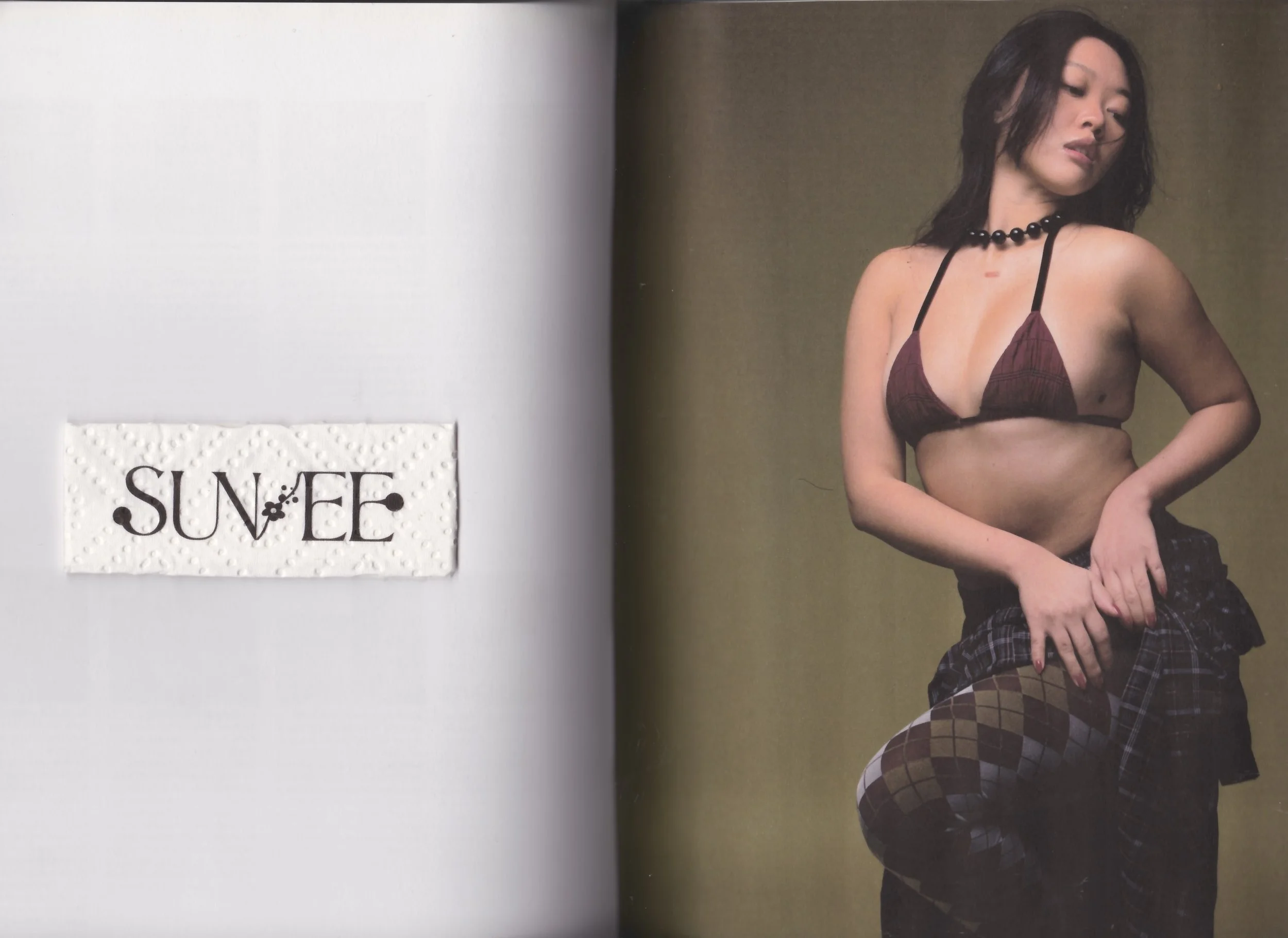 A woman with dark hair wearing a maroon bikini top, black choker, and plaid pants, posing against a plain background.