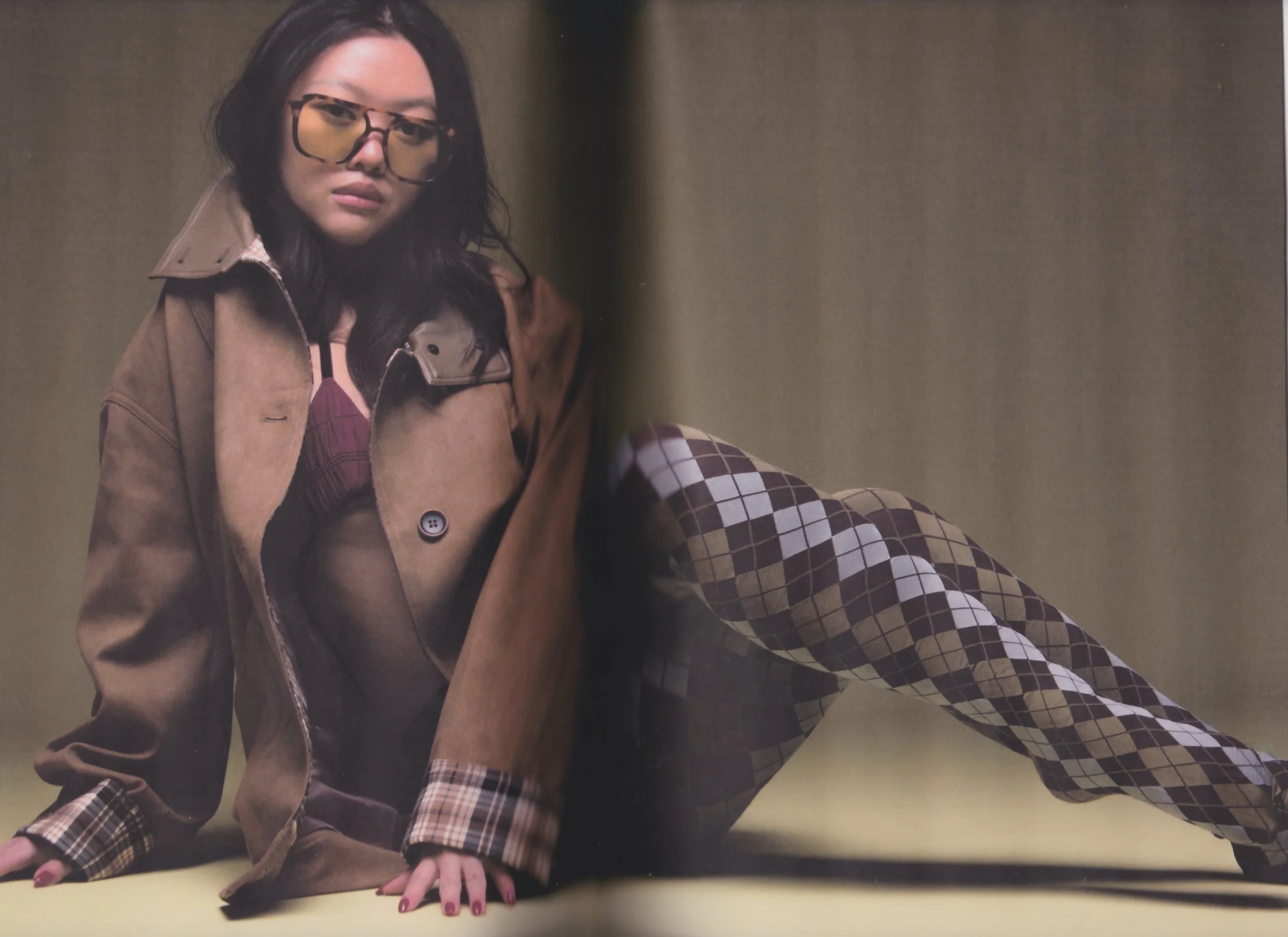A woman with dark hair and glasses posing on a yellow surface against a dark background, wearing a tan coat, a patterned top, and checkered pants.