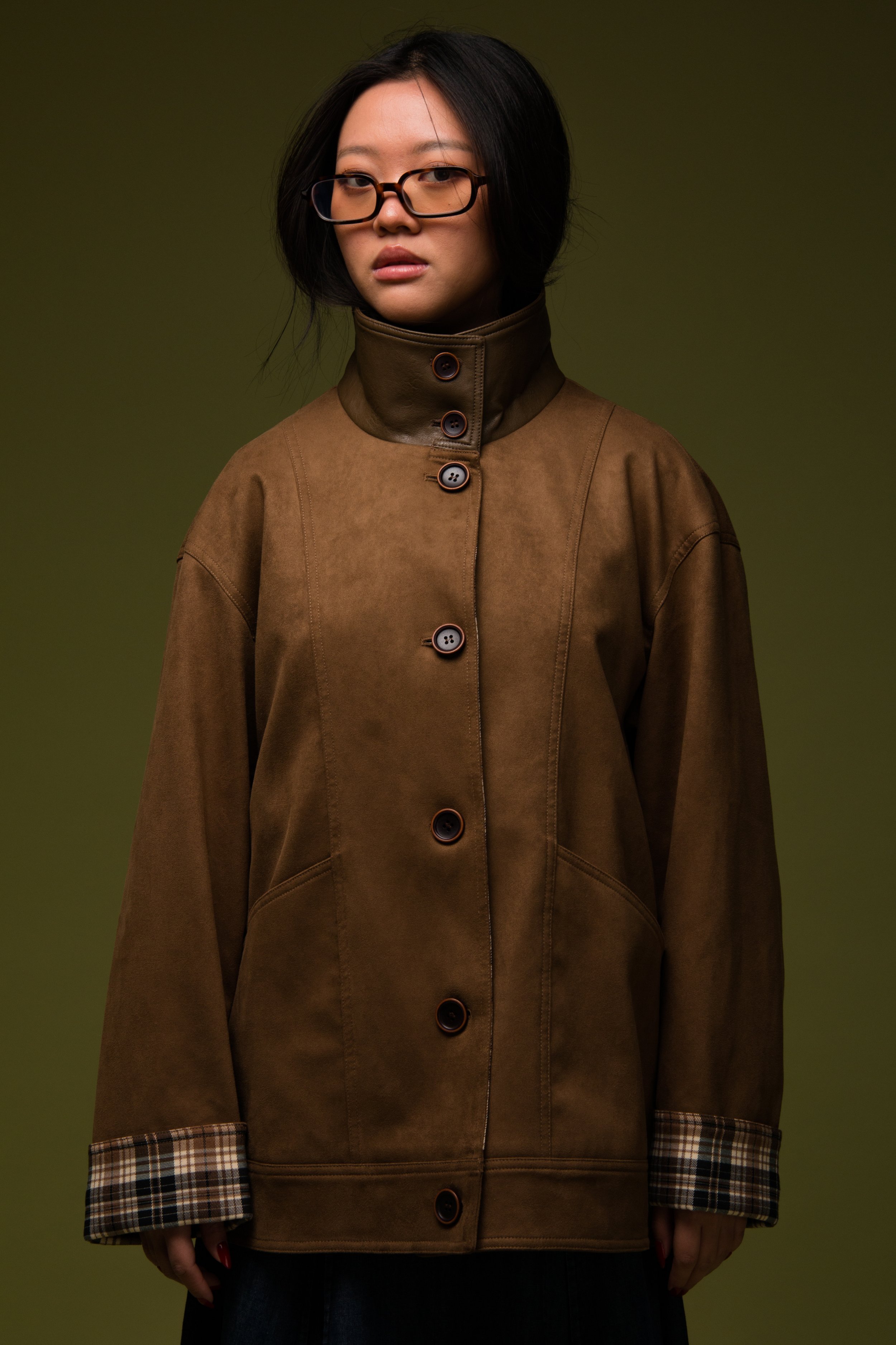 A woman with shoulder-length black hair wearing glasses, a brown button-up coat with a high collar, and plaid cuffs, standing against a plain green background.