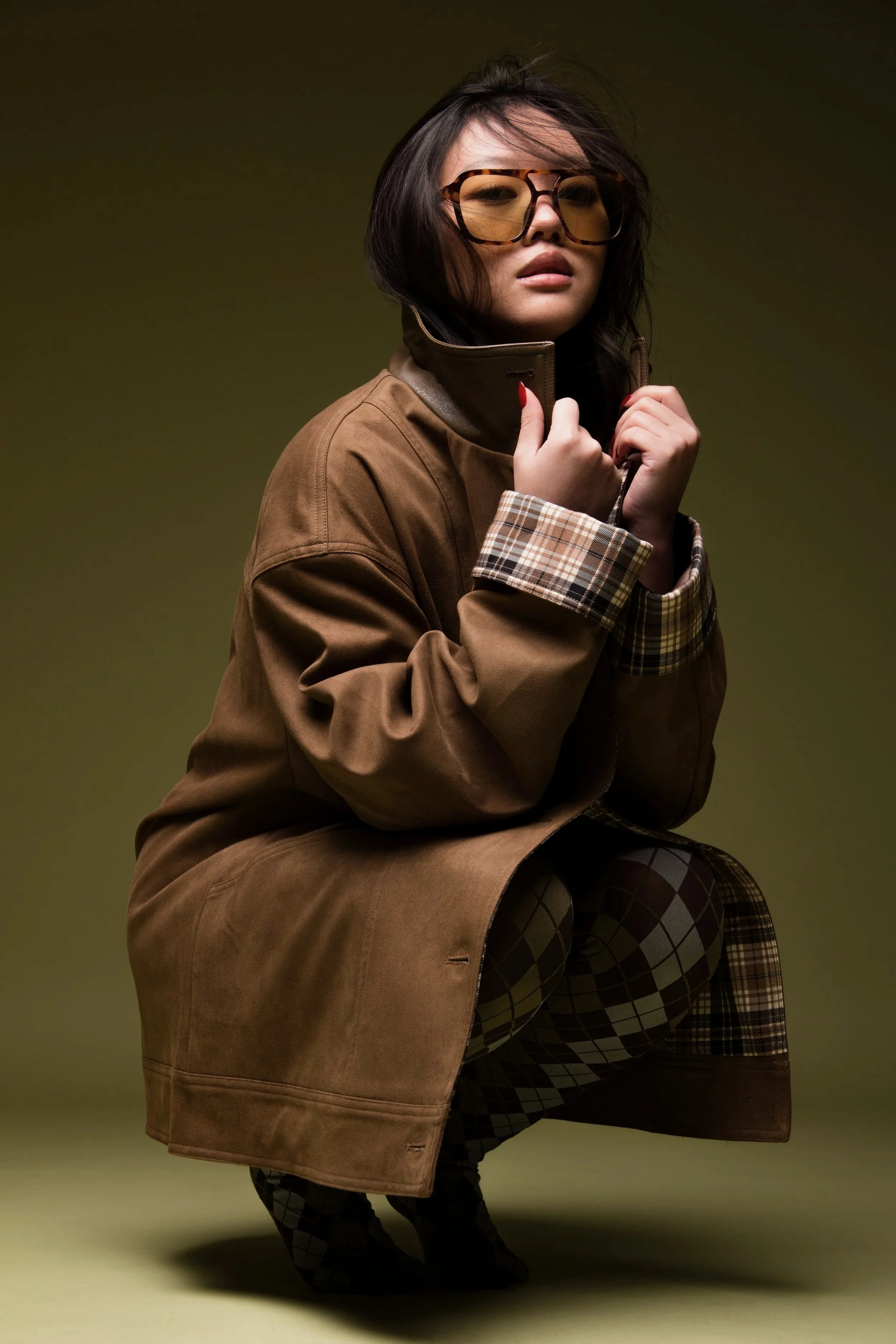 A woman with short dark hair wearing large brown sunglasses, a tan trench coat with plaid checkered cuffs, and patterned pants, sitting in a crouched position against a plain, olive-green background.