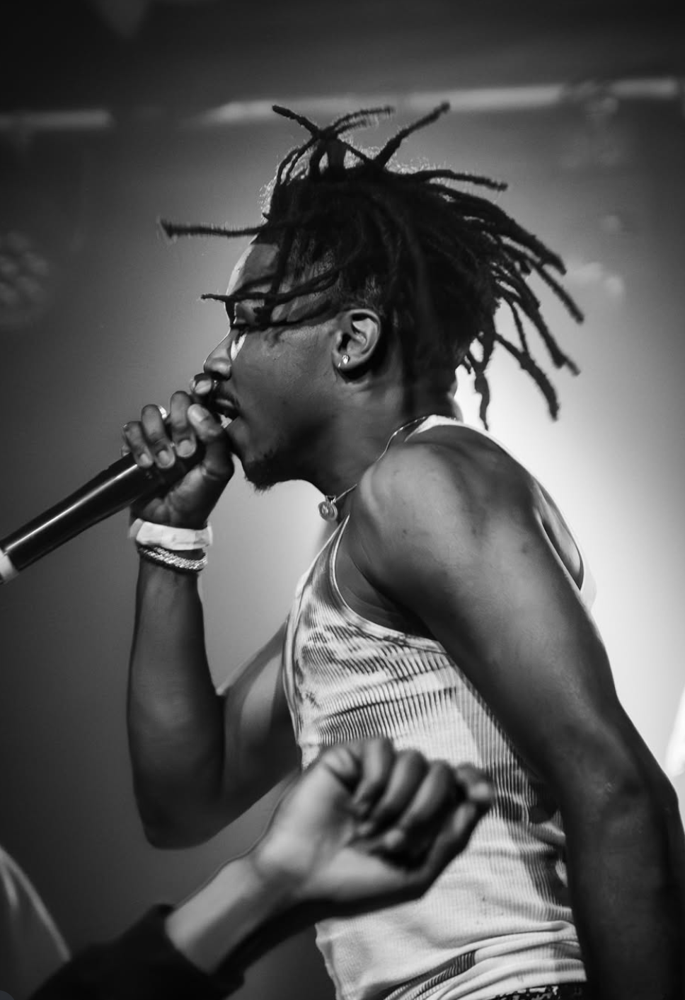 A black and white photo of a person with dreadlocks performing with a microphone, wearing a sleeveless top, earrings, and multiple bracelets.