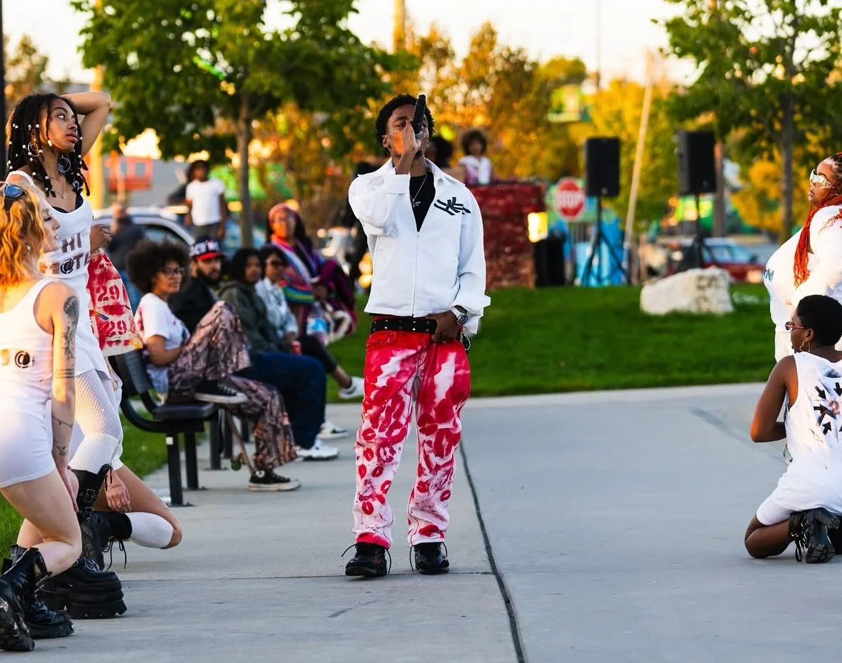 A group of dancers performs outdoors at a park during what appears to be a community event. They are dressed in casual, colorful clothing, with some wearing plaid and tie-dye patterns. An onlooker is taking photos, and there are trees and a stage set