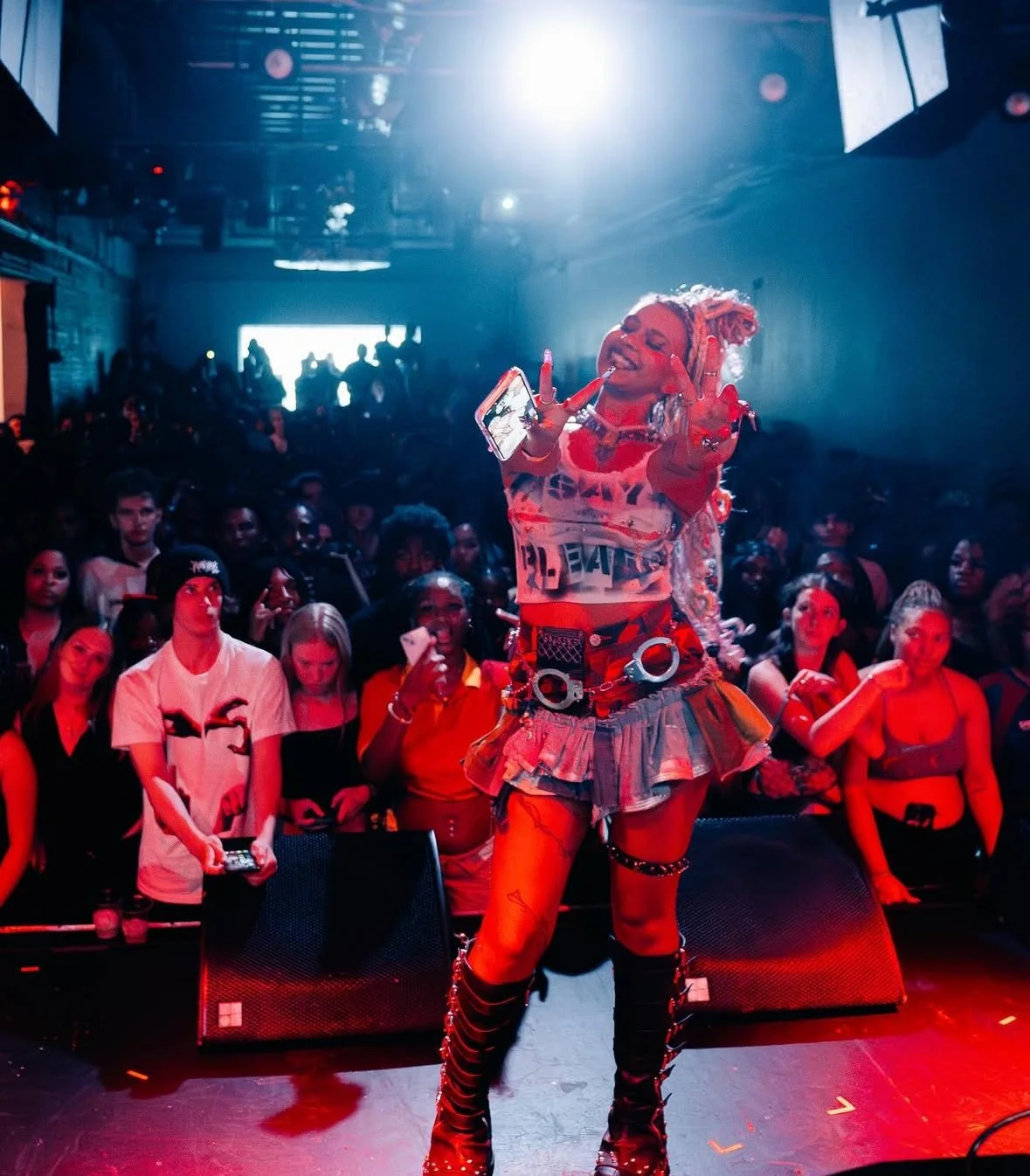 Performer with colorful hair and edgy outfit on stage in front of a large audience at a concert or club.