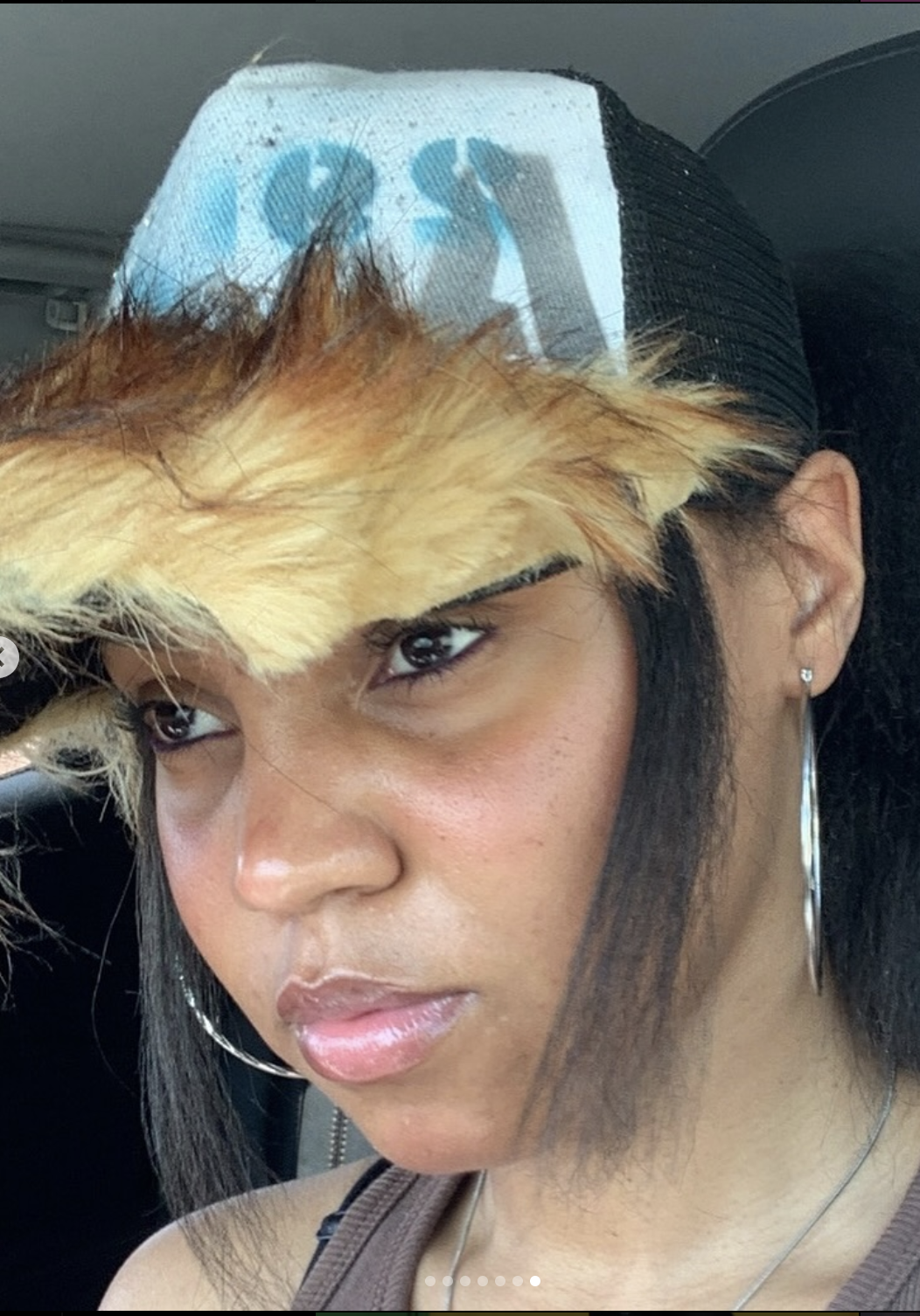 A woman sitting in a car, with a furry, yellowish-orange hat, wearing earrings, with a serious expression.