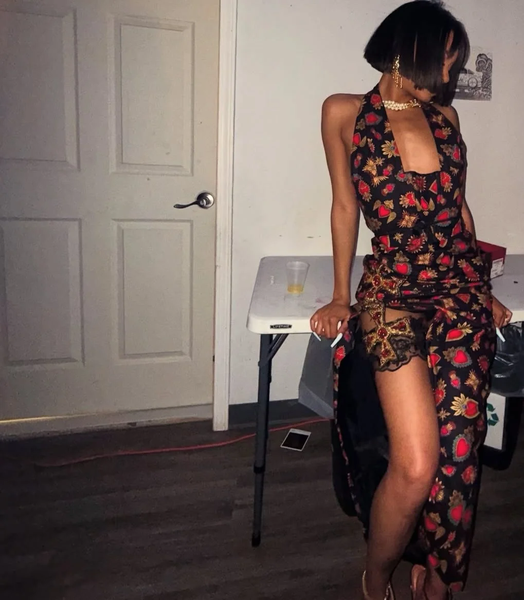 A woman wearing a sheer, floral-patterned dress with lace trim, accessorized with earrings and a pearl necklace, standing by a folding table against a white wall, with a plastic cup and phone on the table.