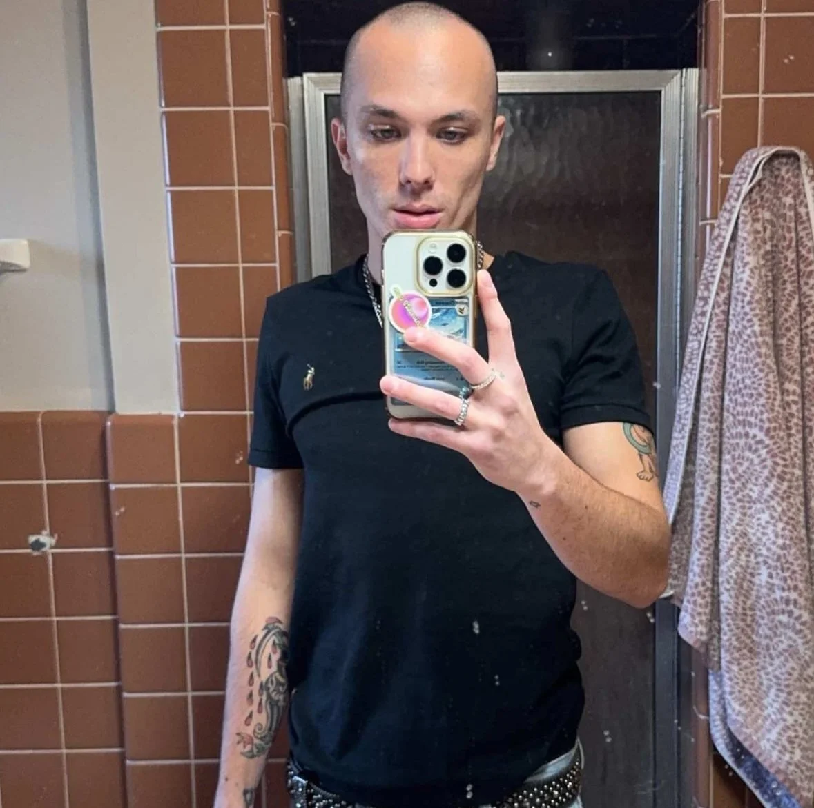 A person taking a mirror selfie in a bathroom, with tattoos visible on their arms, wearing a black shirt with a small logo on it, and holding a phone with a colorful case.
