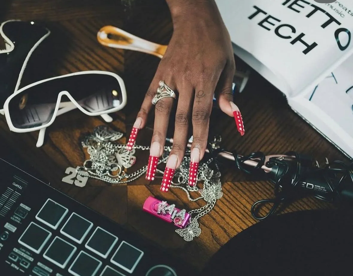 A person's hand with long red nails with white polka dots, wearing a silver ring, resting among jewelry, sunglasses, and electronic devices on a wooden table. Half of a box with the words "METTO TECH" is visible.