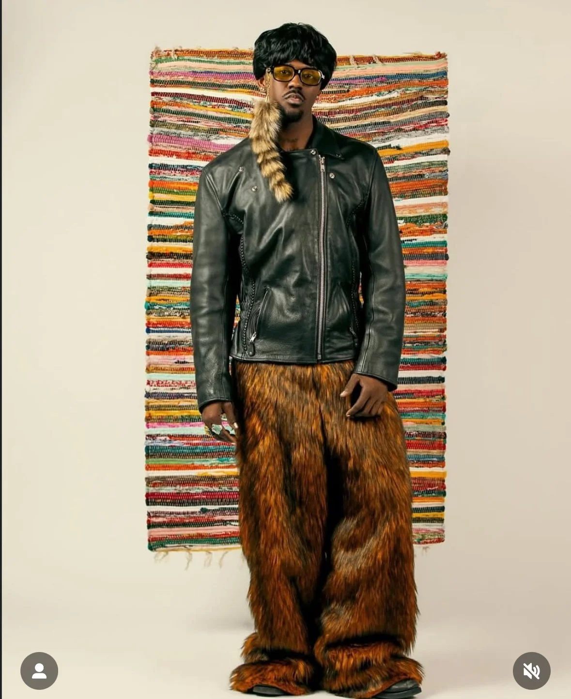 A man dressed in a black leather jacket, furry orange and black pants, and yellow sunglasses stands in front of a colorful woven tapestry.