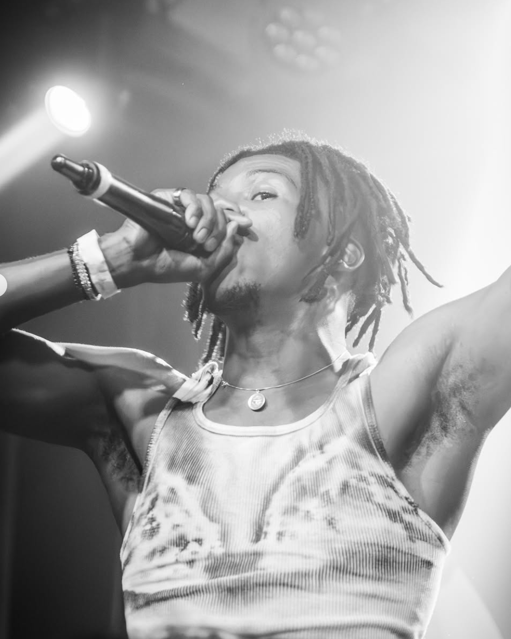 A person with dreadlocks singing into a microphone on stage, wearing a sleeveless shirt and necklace, with stage lighting in the background.