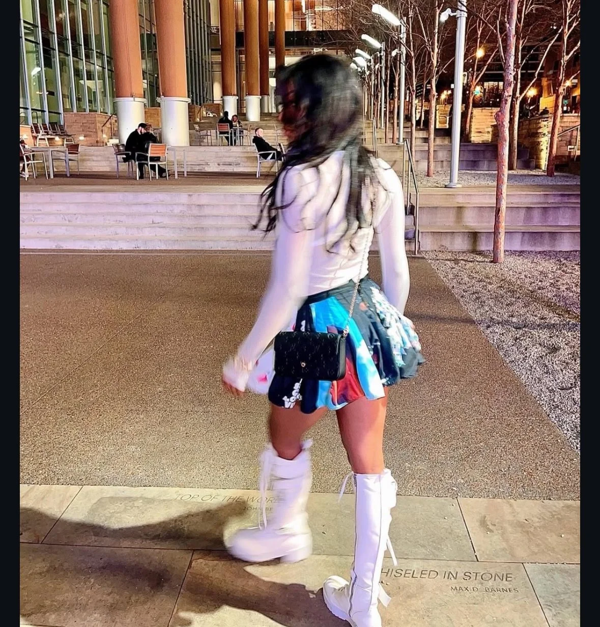 A woman with long hair wearing a white top, colorful shorts, and white thigh-high boots standing on a sidewalk at night in front of a modern building and outdoor seating area with trees and streetlights.