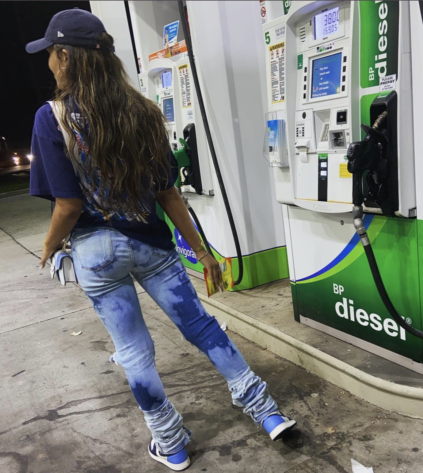 A woman in a blue baseball cap, a blue T-shirt, and ripped jeans is refueling her vehicle with diesel at a BP gas station during nighttime. She is holding a debit or credit card and standing in front of the fuel pump.