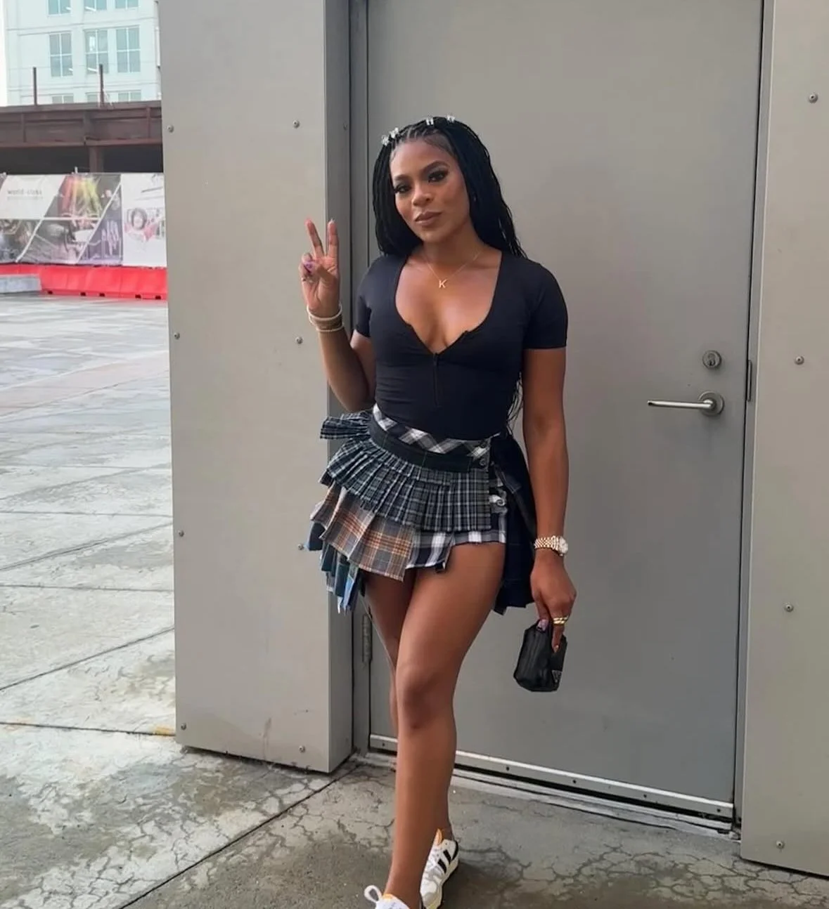 A woman with long dark hair in a black top and a multi-layered plaid skirt, making a peace sign with her right hand, standing outdoors in front of a gray door, holding a small black purse in her left hand.