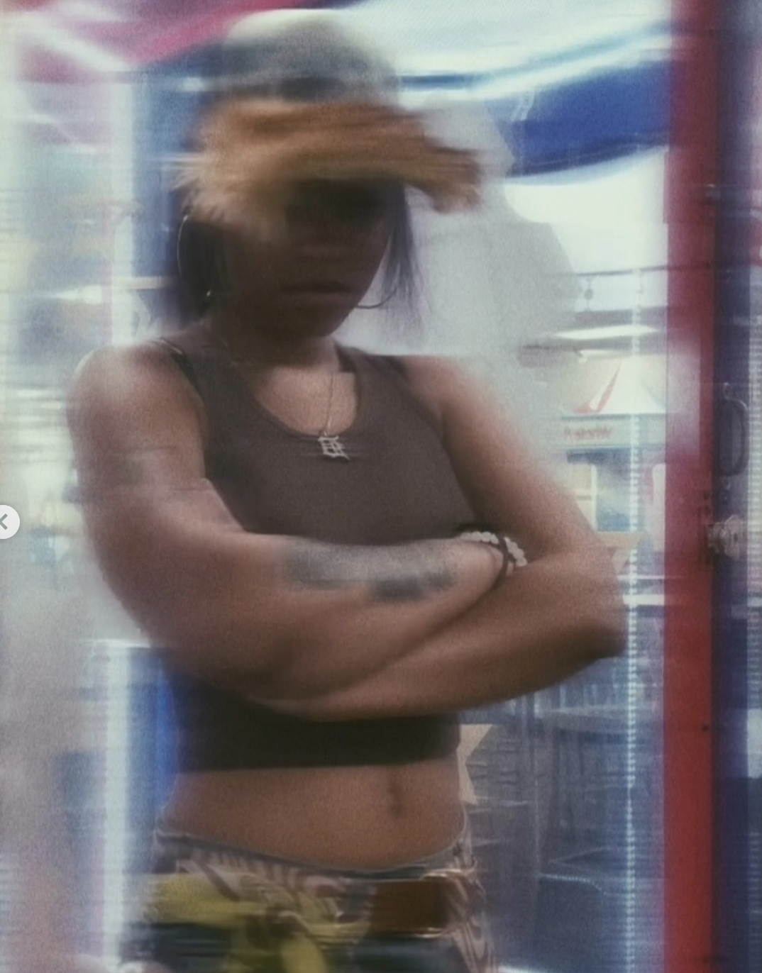 A woman with a blurred face, wearing a black crop top, standing with arms crossed in front of a store display, with a yellow item partially visible at the bottom.