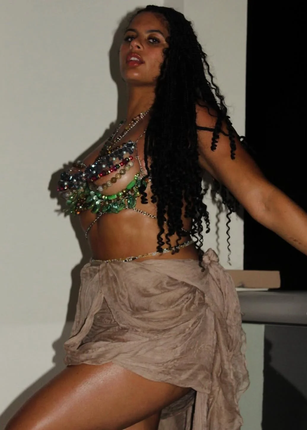 A woman with long curly black hair, wearing multiple colorful bead necklaces, poses confidently against a neutral background.