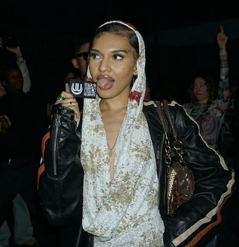 A young woman with a headscarf sticking out her tongue while holding a phone with a QR code, standing in a crowd indoors.