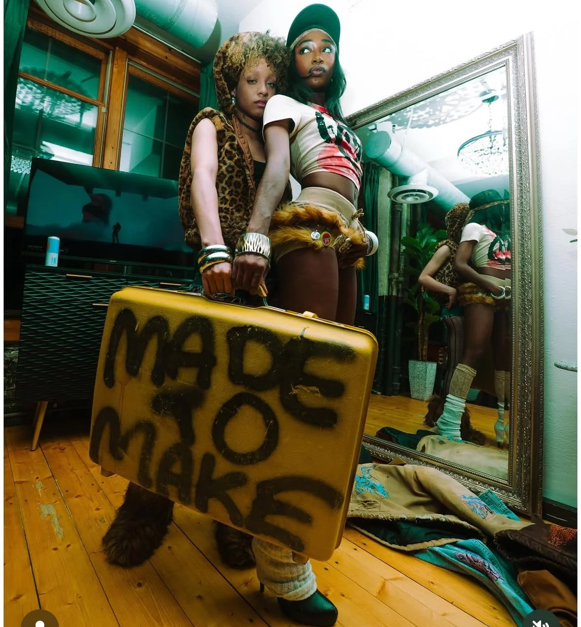Two women in colorful, edgy outfits taking a mirror selfie in a room, one holding a yellow suitcase with black graffiti writing and wearing furry boots.