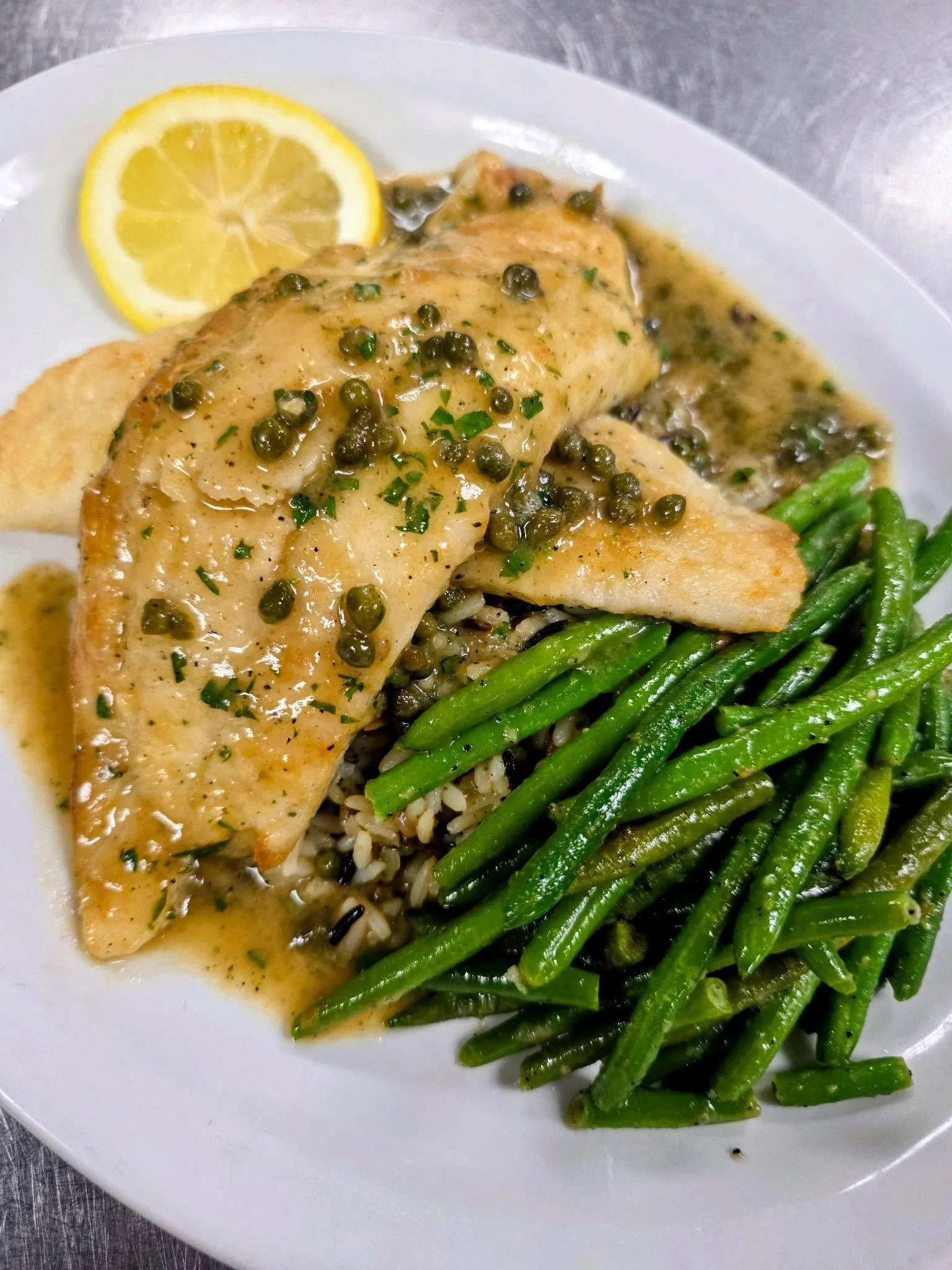 A plate of cooked fish topped with green peppercorn sauce, served with lemon slices, green beans, and rice.