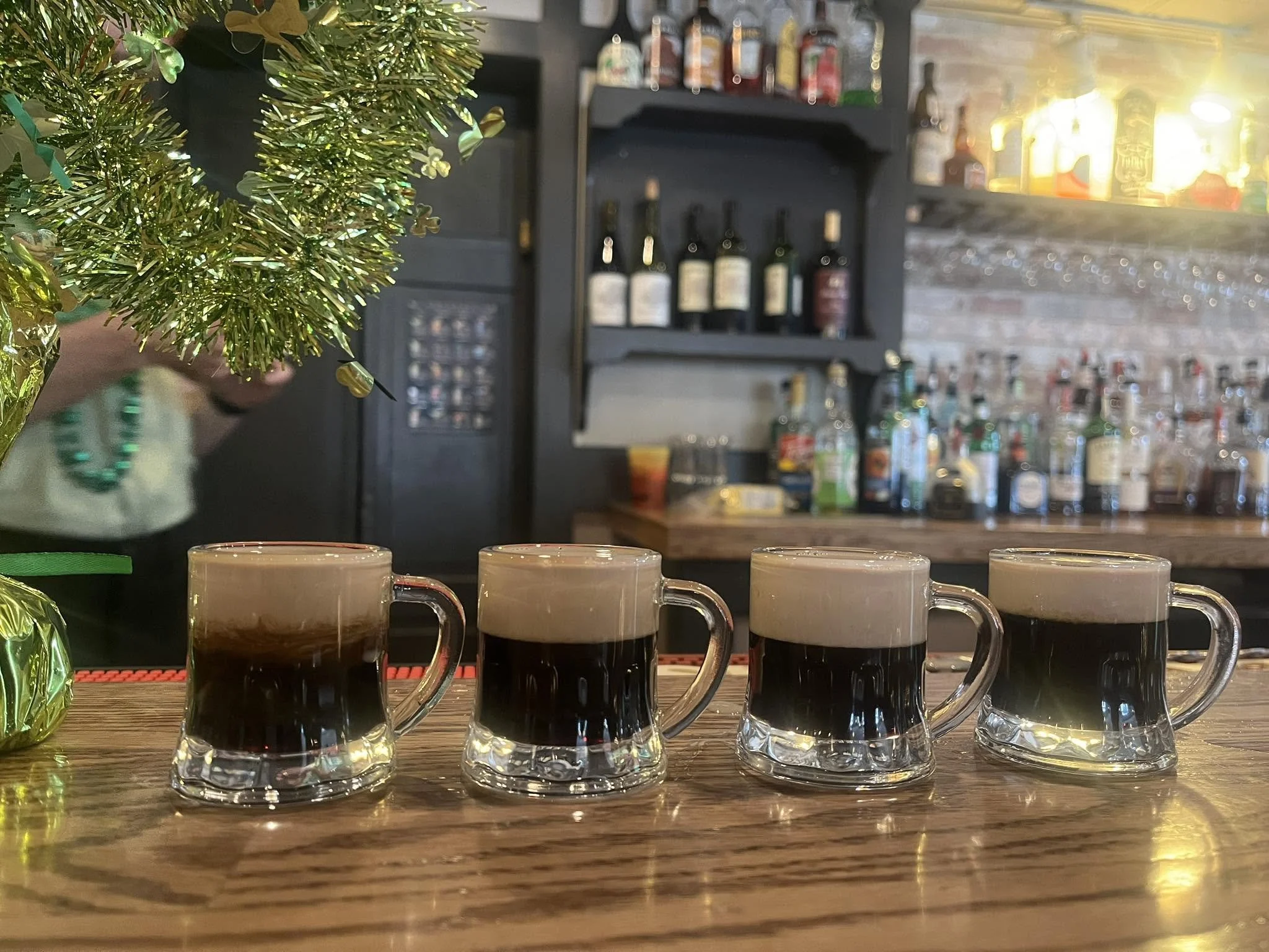 A wooden bar counter with six glass coffee mugs filled with black coffee lined up in a row. Behind the counter, there is a small decorative Christmas tree with green and gold ornaments, and a bar shelving unit stocked with various bottles and jars, i