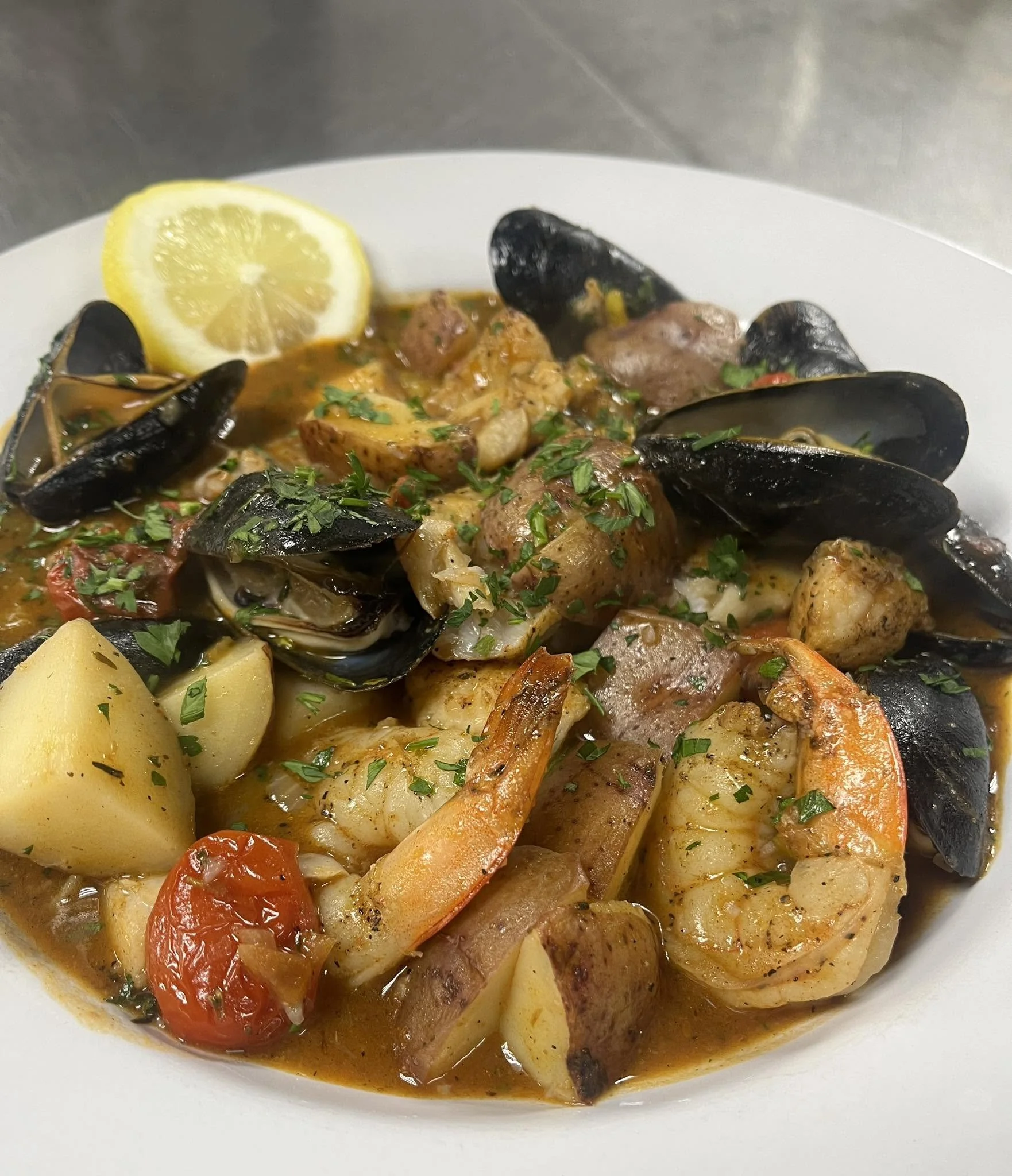Seafood stew with mussels, shrimp, and fish pieces garnished with chopped parsley, served with lemon and potatoes.