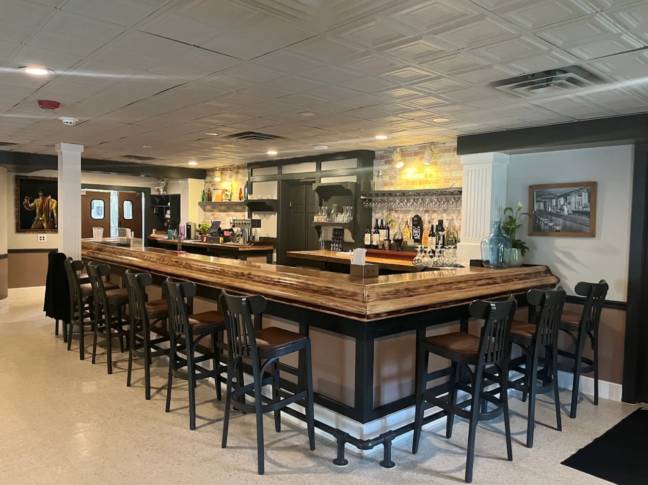 A cozy bar with a large, U-shaped wooden counter and black chairs, various bottles and glasses behind the bar, a painting of a performer on the left wall, and a framed black-and-white photo on the right wall.