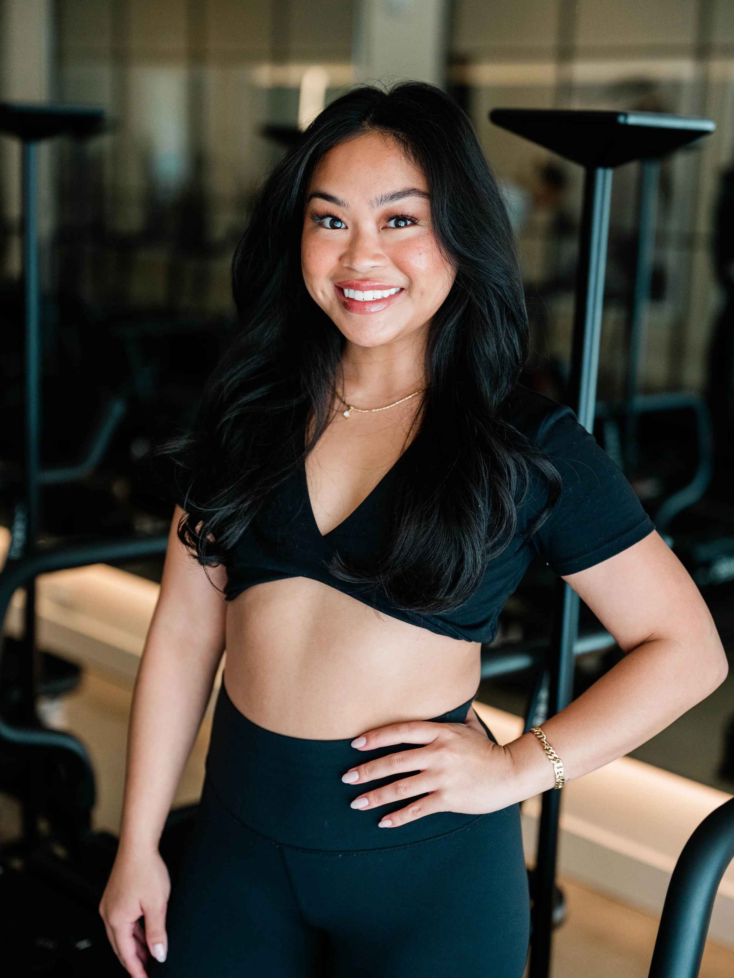 A woman with long black hair and makeup is smiling at the camera. She is wearing a black crop top and black leggings, standing indoors in a gym or fitness studio.