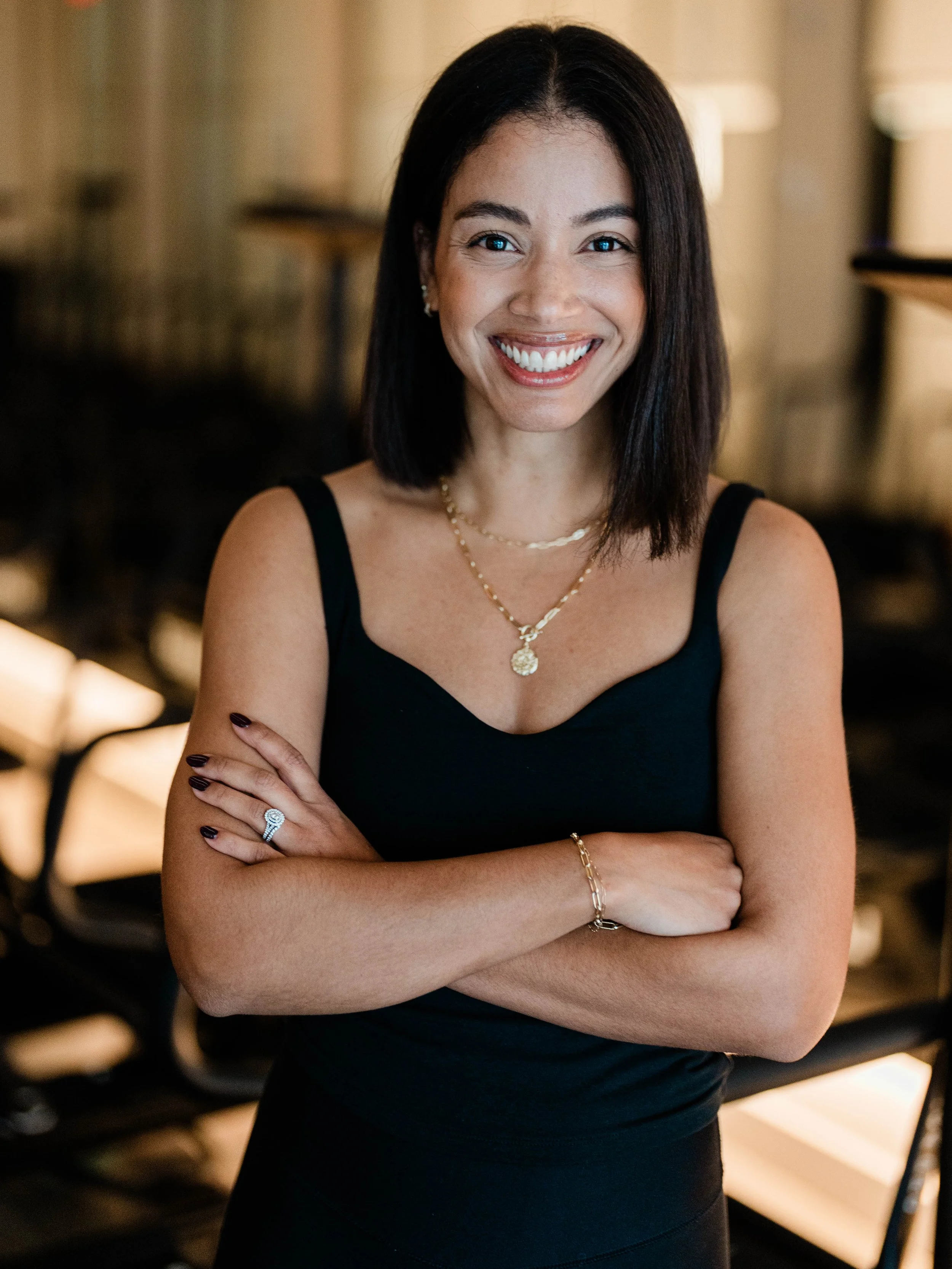 A woman with shoulder-length black hair, smiling, wearing a black sleeveless top, gold necklaces, a ring, and a bracelet, standing arms crossed in an indoor setting with warm lighting.