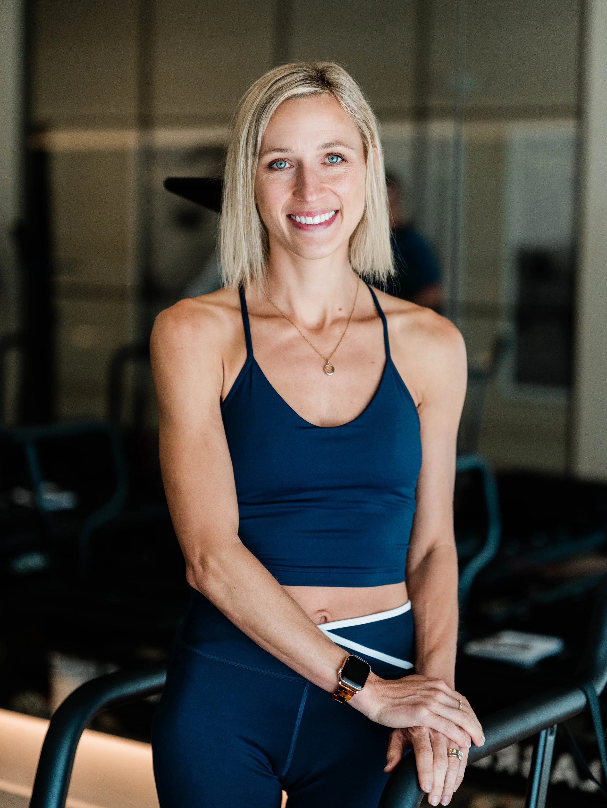 A smiling woman with blonde hair, wearing a navy blue athletic tank top and matching leggings, standing in a gym environment.