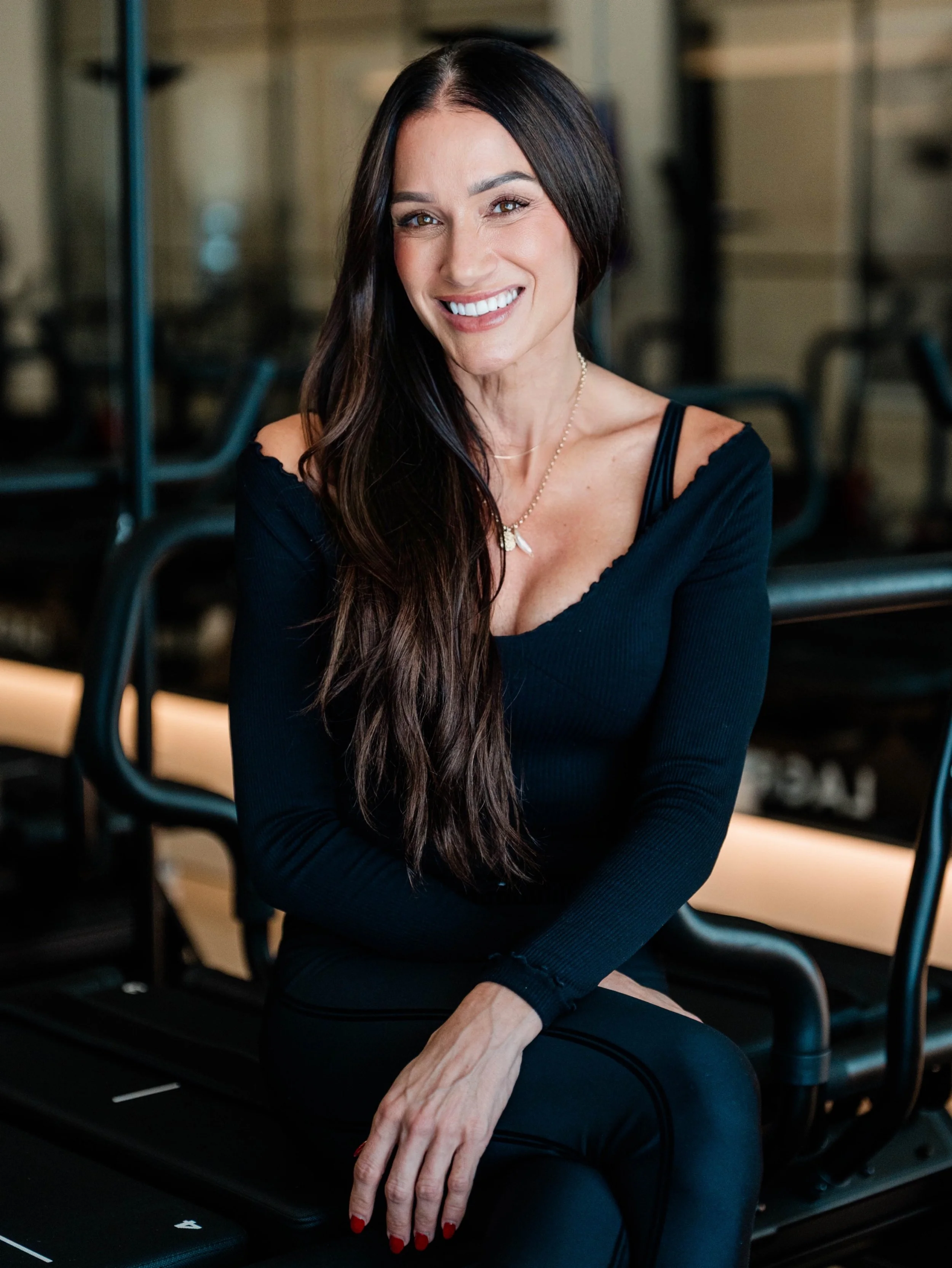 Amy is a lagree instructor - she is pictured wearing a black off-shoulder top with a necklace, seated in a gym with workout equipment in the background.