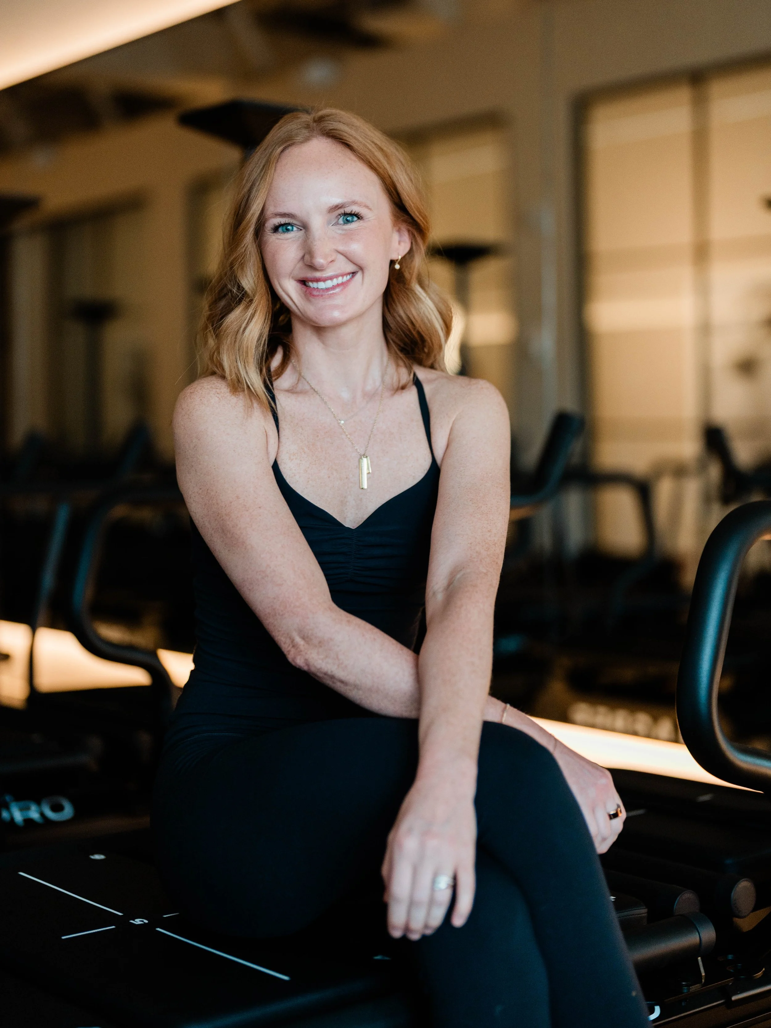 Maddie teaches Lagree, VersaClimber and Yoga - She is pictured sitting in a room with warm lighting and gym equipment in the background.